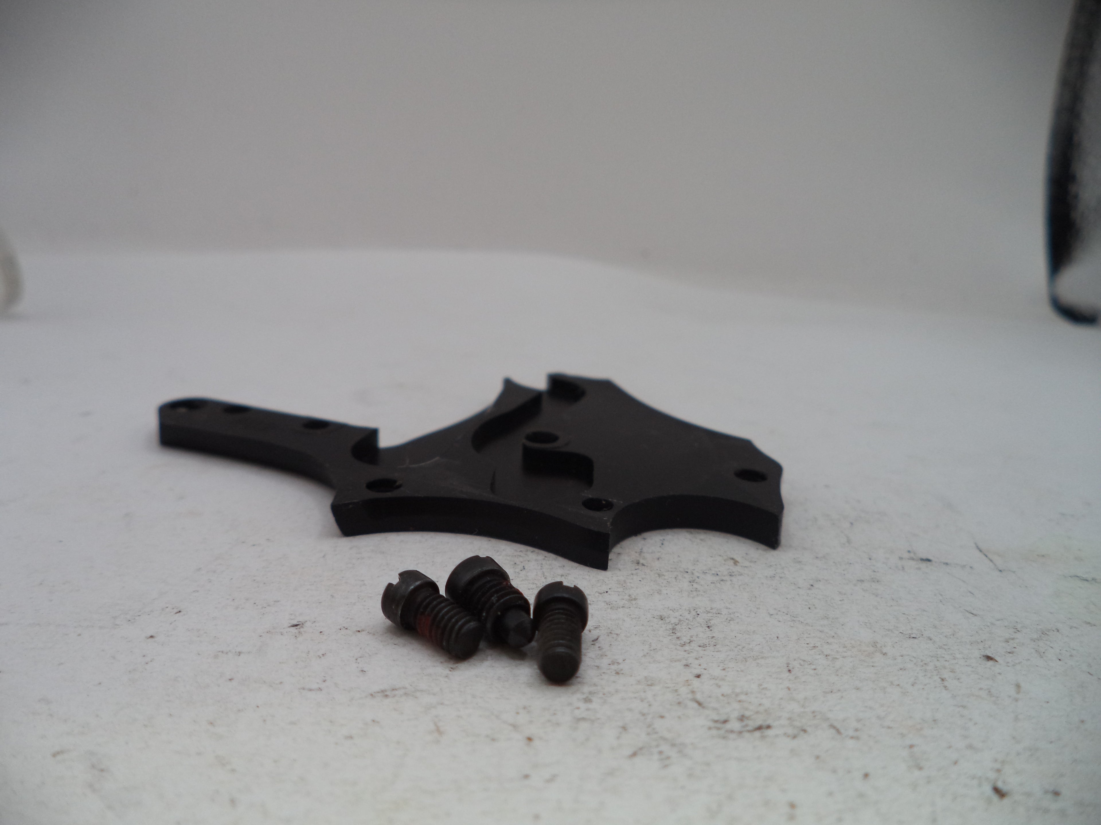 A black plastic mounting bracket with three small black screws, photographed on a light gray surface.