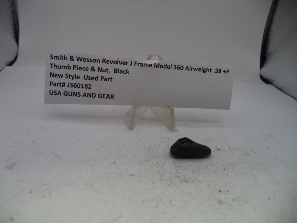 A small black used replacement part, identified as a Smith & Wesson Revolver J Frame thumb piece and nut, displayed on a clear plastic stand next to a white paper label card on a white surface.