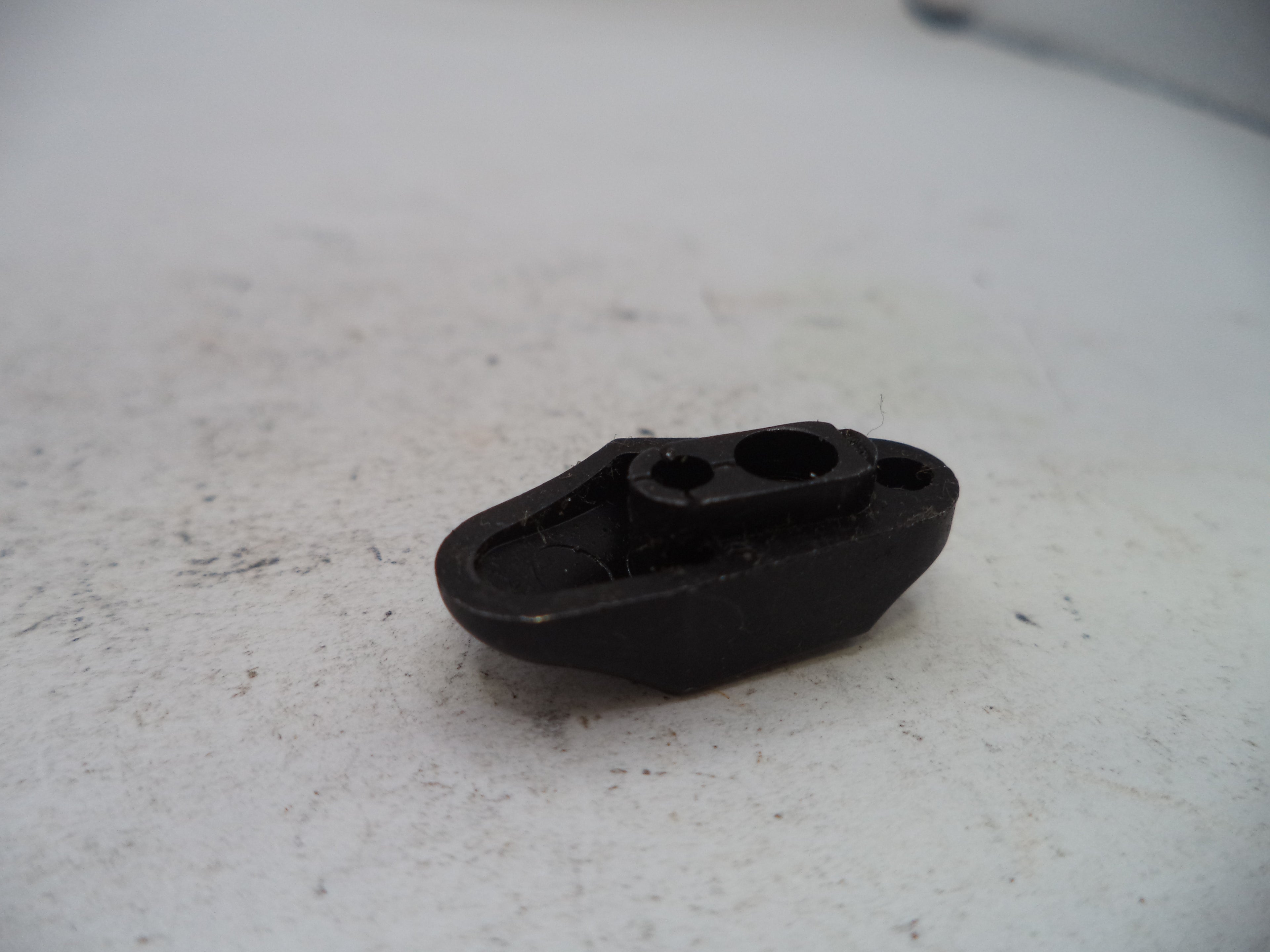 A small black plastic mechanical part with two circular openings, resting on a light gray textured surface.