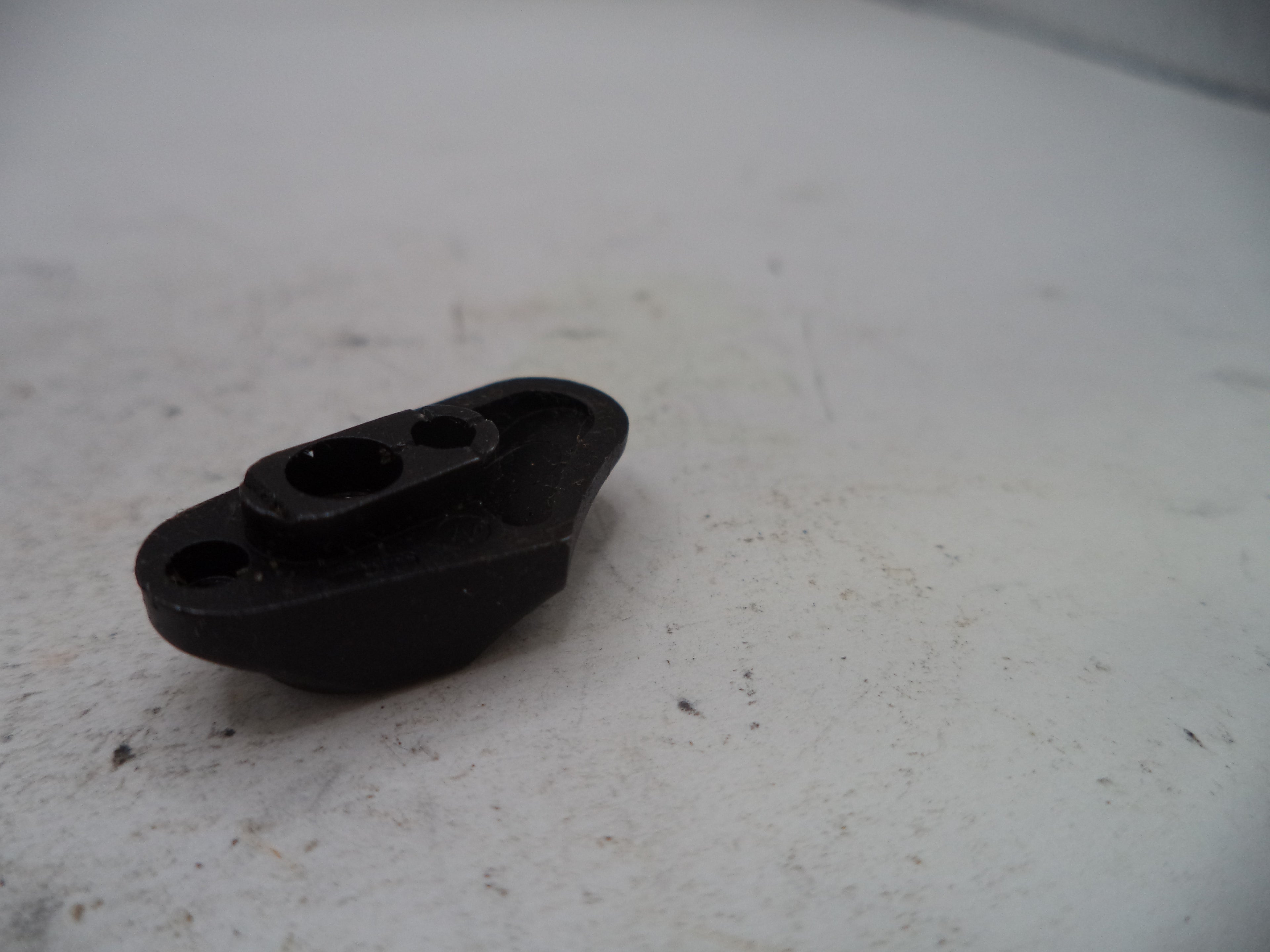 A small black plastic mechanical or automotive part with two circular holes, resting on a dirty, off-white surface.