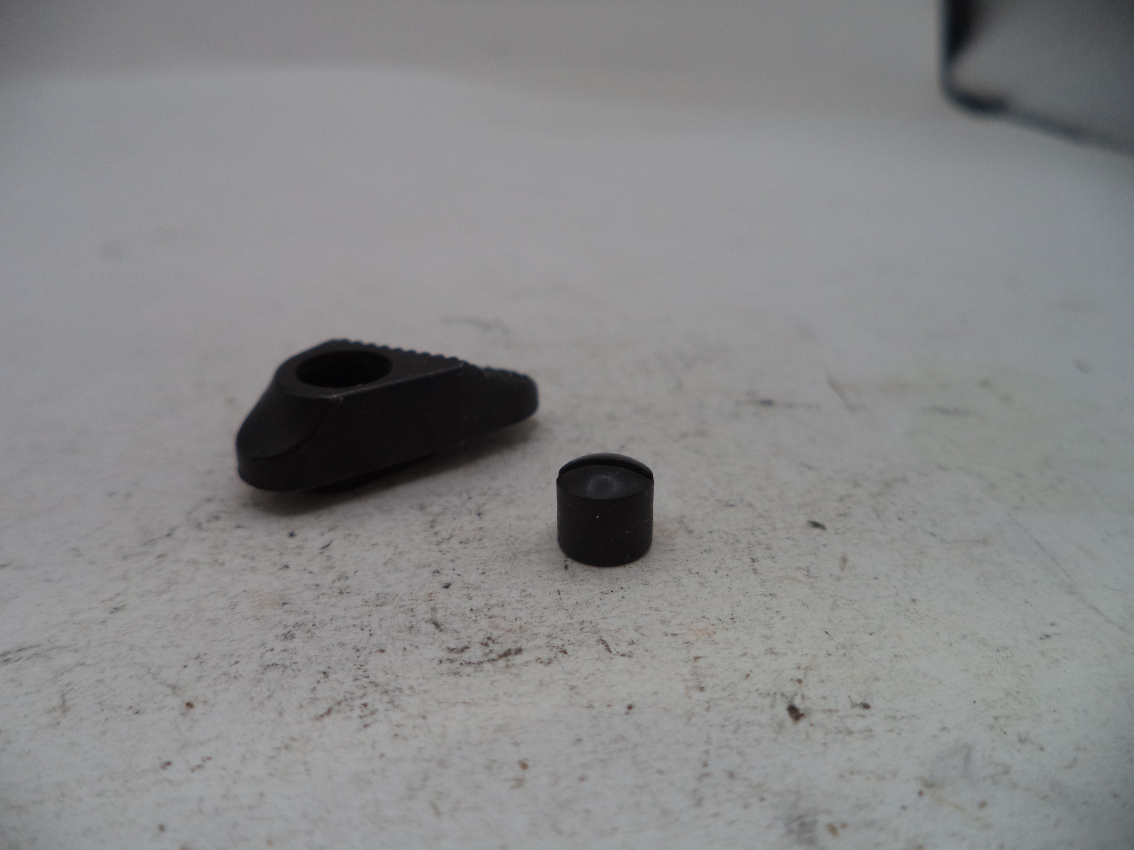 A small black plastic or metal hardware component with a triangular shape and a hole, shown next to a small cylindrical black piece on a light gray textured surface.