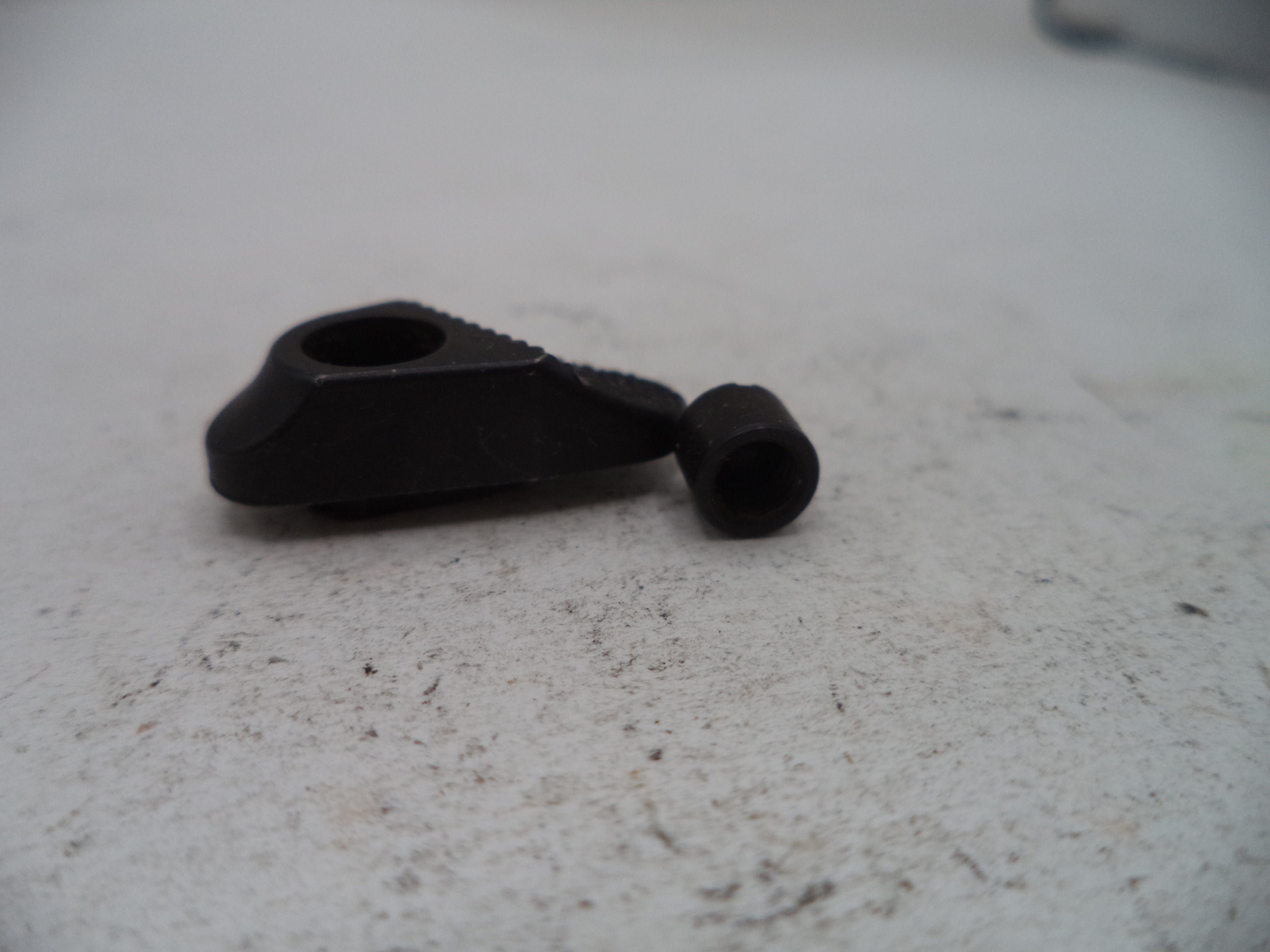 A small black plastic or metal hardware component with a triangular shape and a cylindrical hole, shown next to a matching small black cylindrical piece on a light gray textured surface.