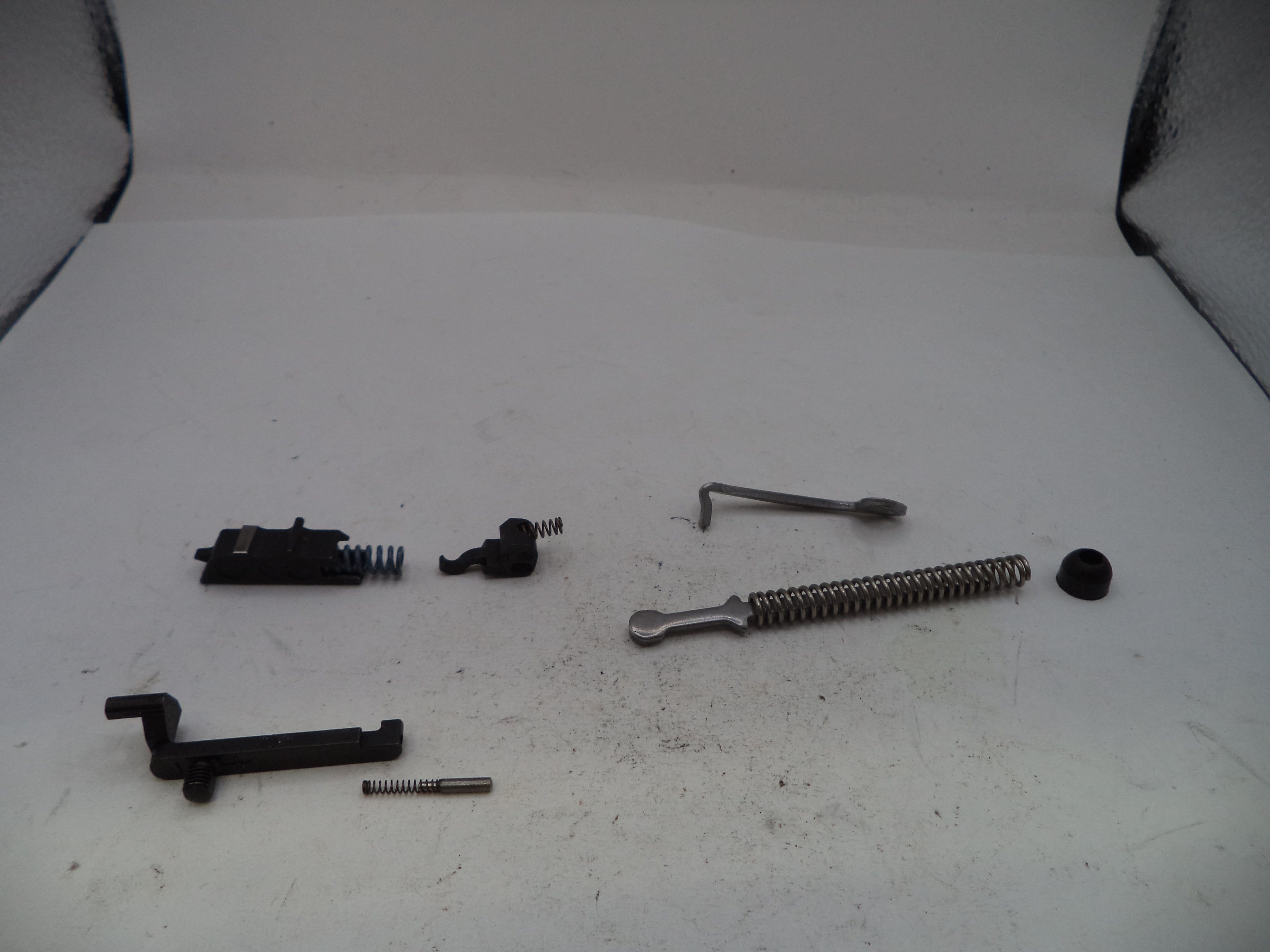 A set of disassembled metal and black mechanical parts, including springs, rods, and small hardware pieces, arranged on a white surface.