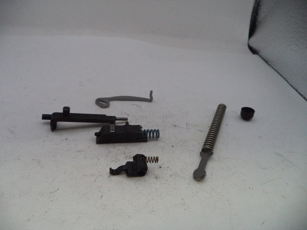 A disassembled black metal firearm component set, including a trigger assembly, recoil spring, safety lever, and other small metal parts, arranged on a white surface.