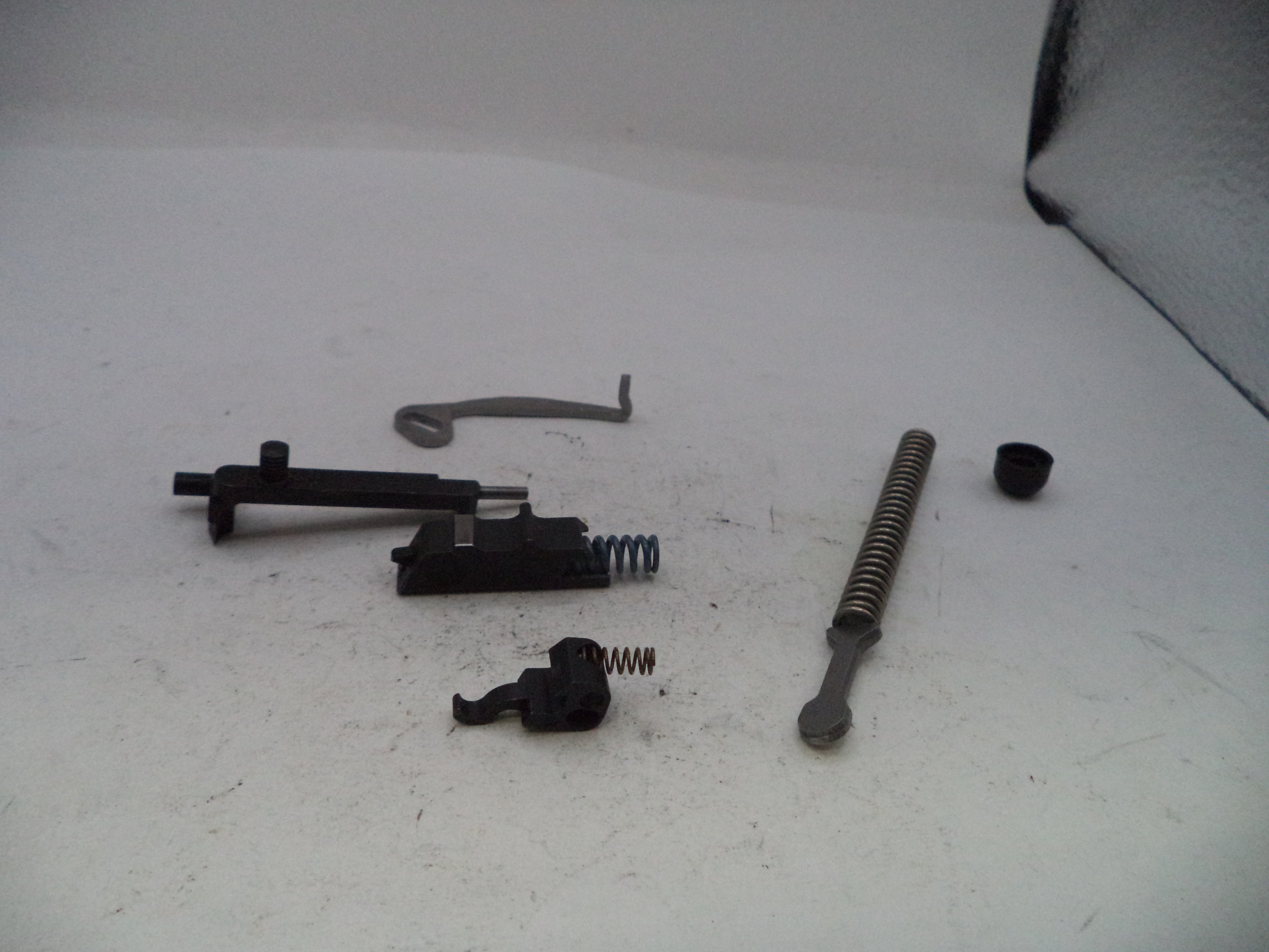 A disassembled black metal firearm component set, including a trigger assembly, recoil spring, safety lever, and other small metal parts, arranged on a white surface.