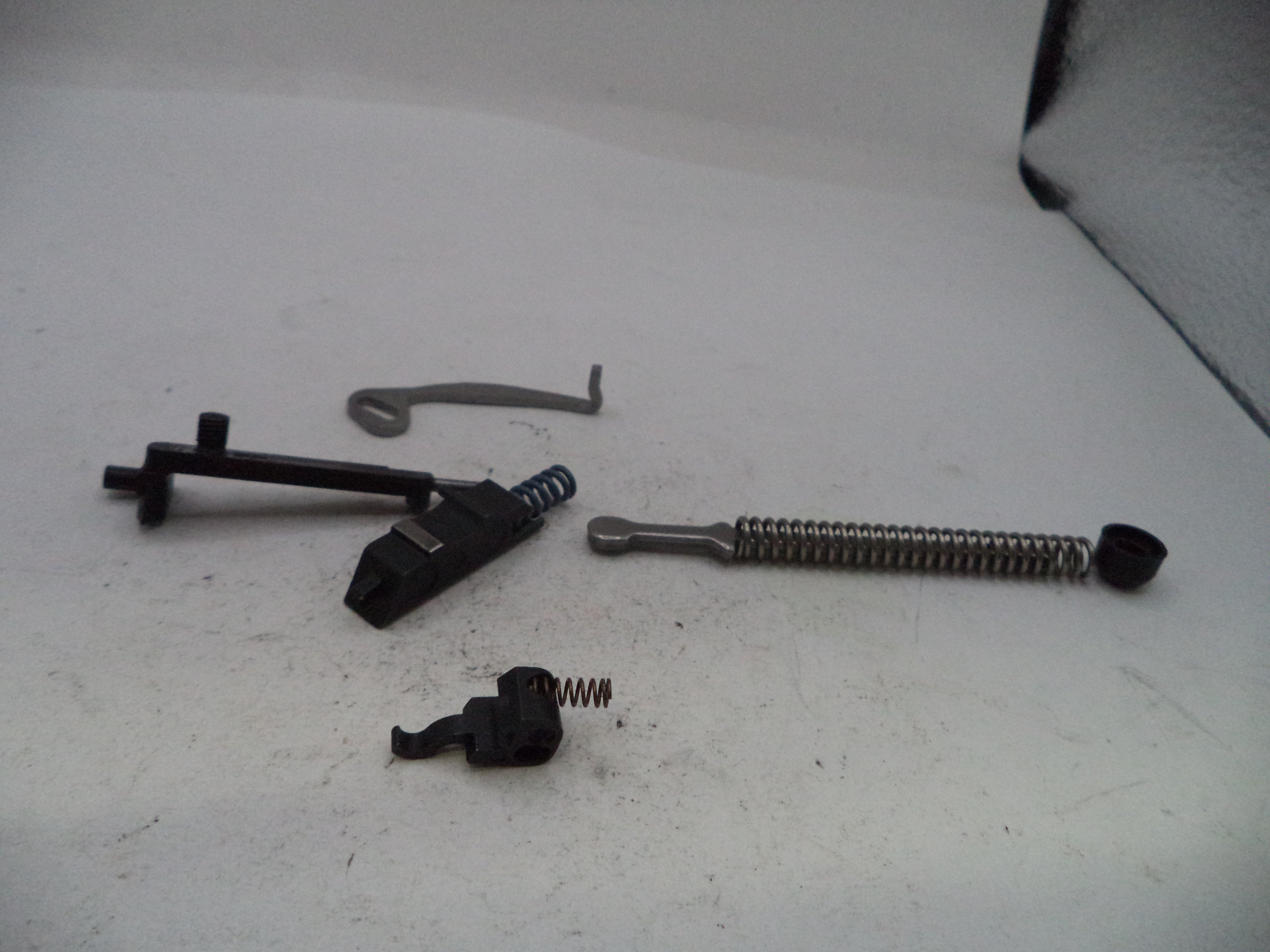 A set of firearm replacement parts including a black trigger assembly, a metal coil spring, a small black spring, and a curved metal lever, laid out on a white surface.