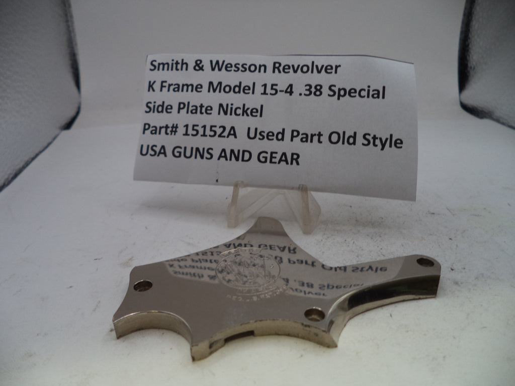 A used metal side plate for a Smith & Wesson revolver, displayed on a clear plastic stand with a paper label card behind it on a white surface.