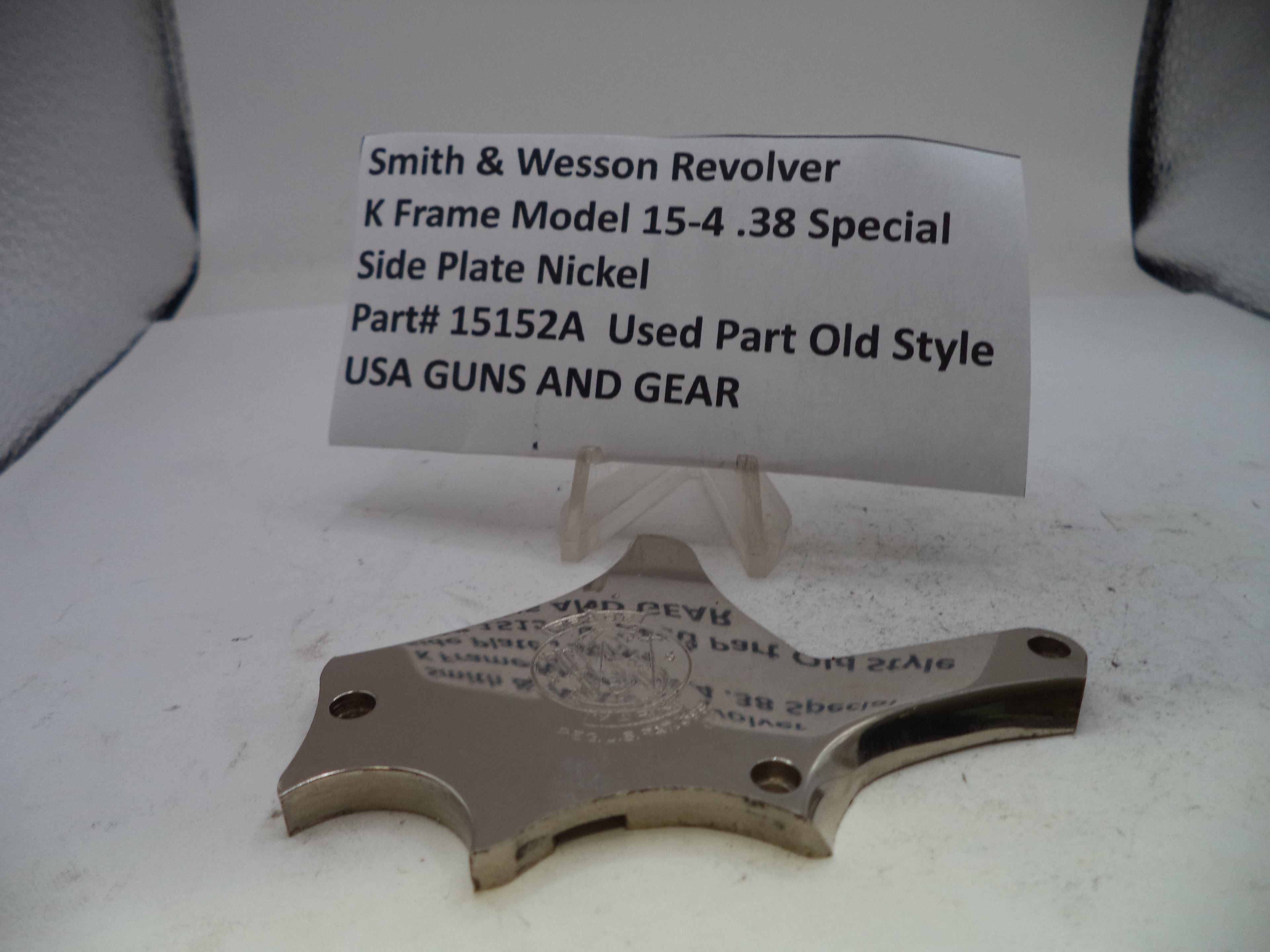 A used metal side plate for a Smith & Wesson revolver, displayed on a clear plastic stand with a paper label card behind it on a white surface.