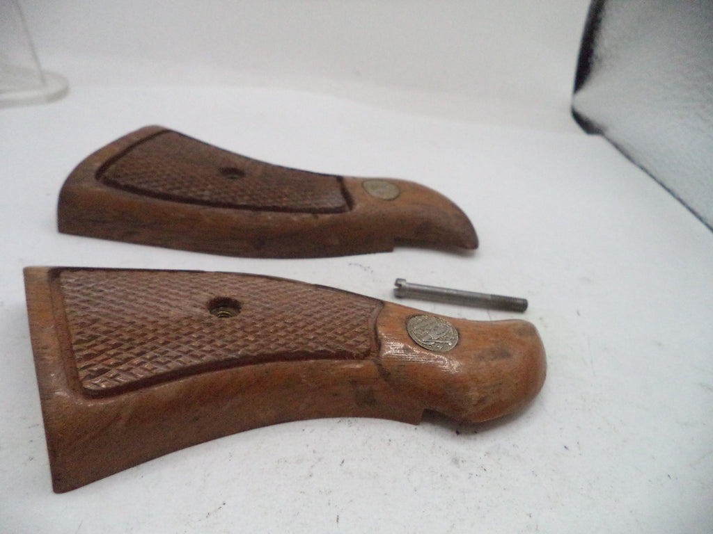 Two brown wooden gunstock grips with textured grip panels and metal medallions, shown on a white surface with a small metal tool between them.
