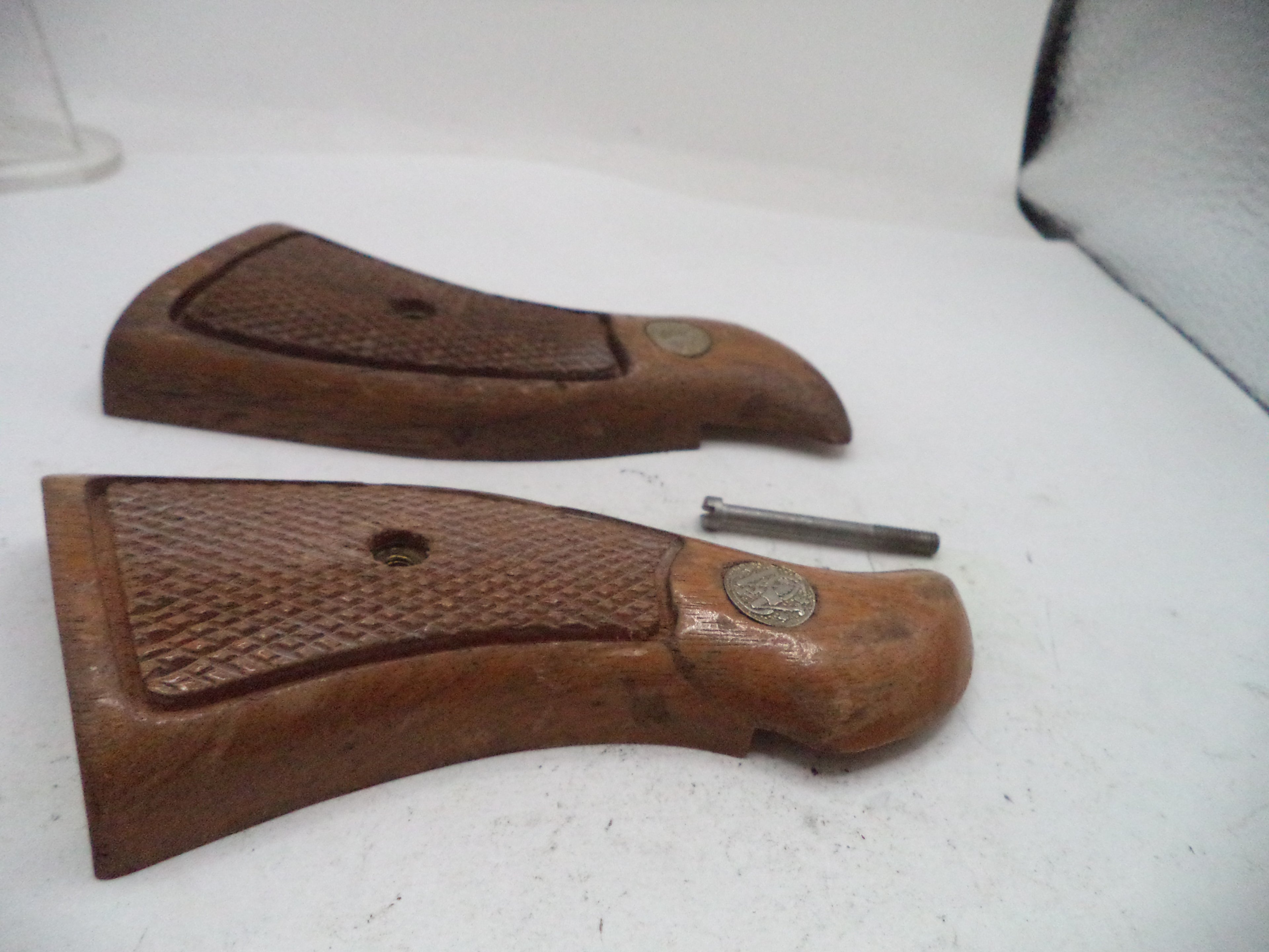Two brown wooden gunstock grips with textured grip panels and metal medallions, shown on a white surface with a small metal tool between them.
