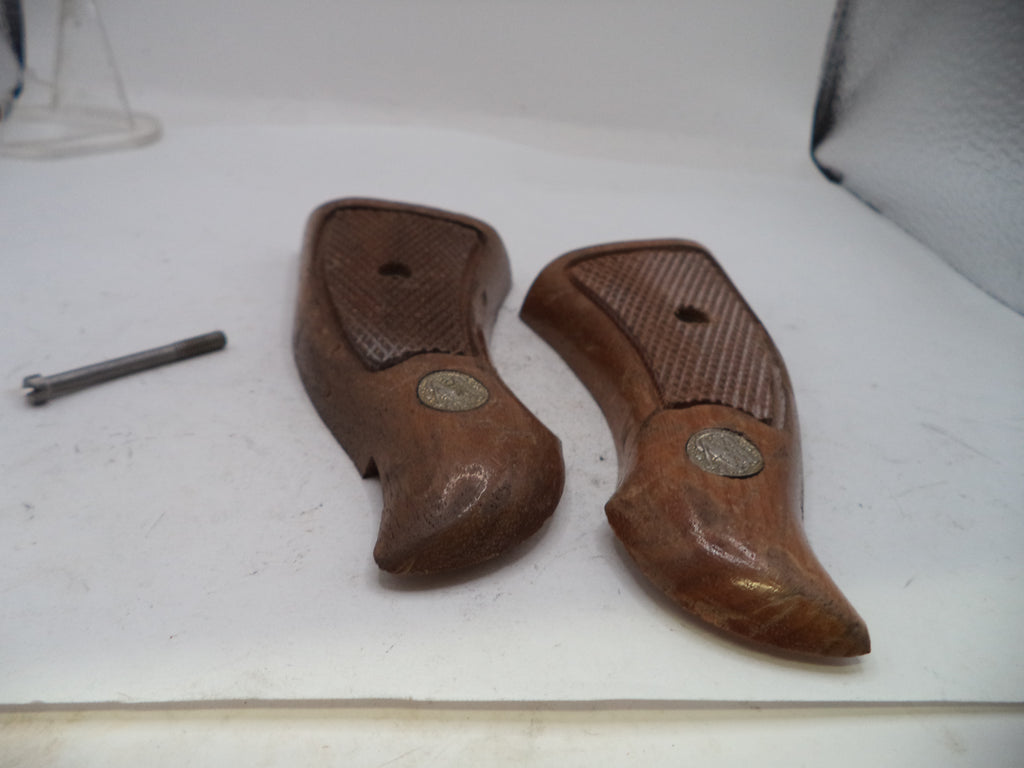 A pair of brown wooden gunstock grips with a textured grip pattern and a small round metal emblem, shown on a white surface next to a small metal screw.