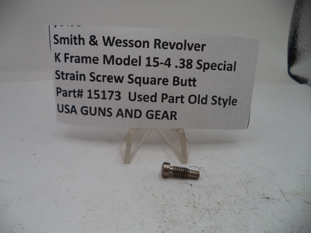 A small metal revolver strain screw part displayed on a clear plastic stand next to a paper label card that reads 'Smith & Wesson Revolver K Frame Model 15-4 .38 Special Strain Screw Square Butt Part# 15173 Used Part Old Style USA GUNS AND GEAR'.