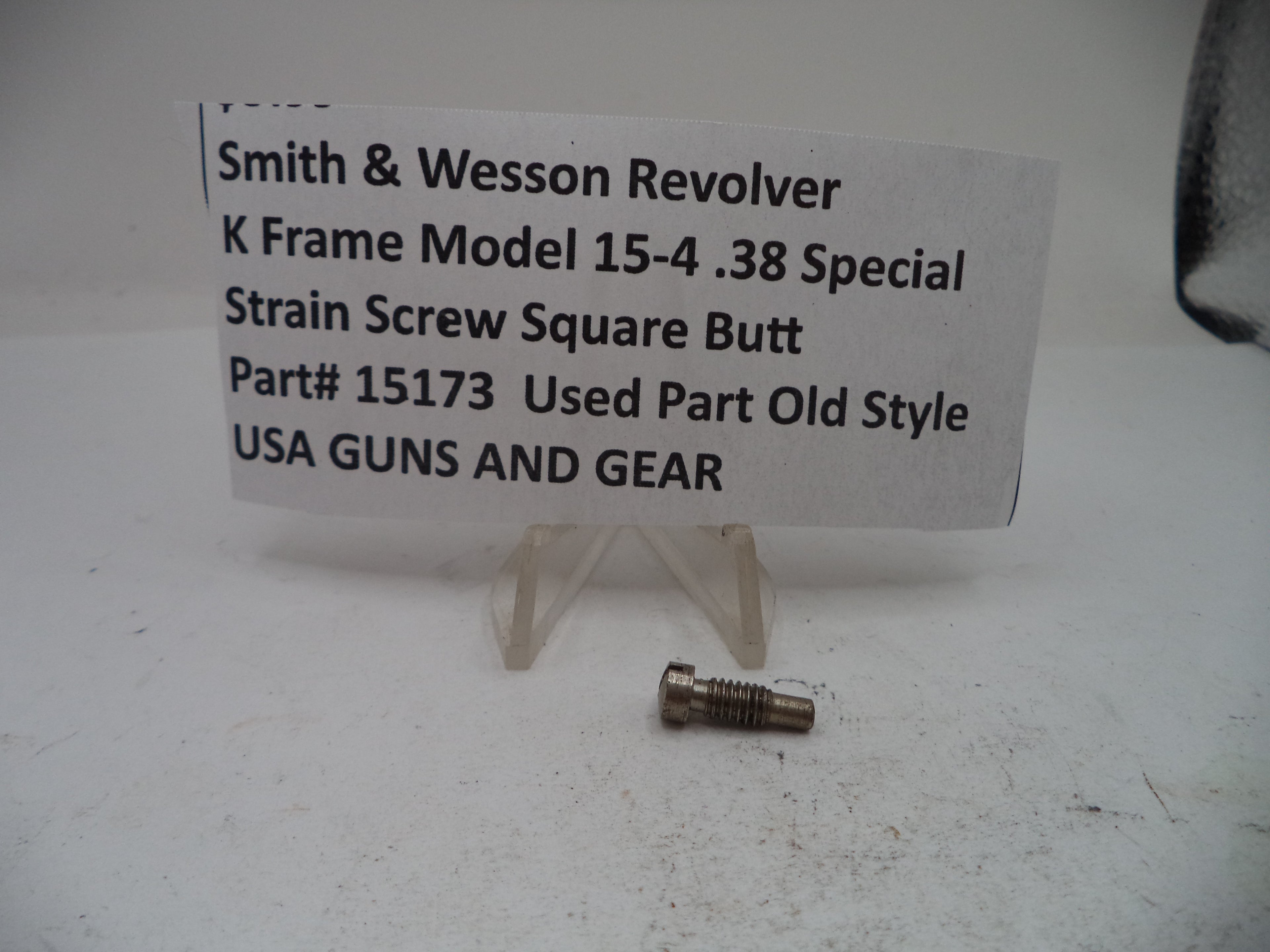 A small metal revolver strain screw part displayed on a clear plastic stand next to a paper label card that reads 'Smith & Wesson Revolver K Frame Model 15-4 .38 Special Strain Screw Square Butt Part# 15173 Used Part Old Style USA GUNS AND GEAR'.