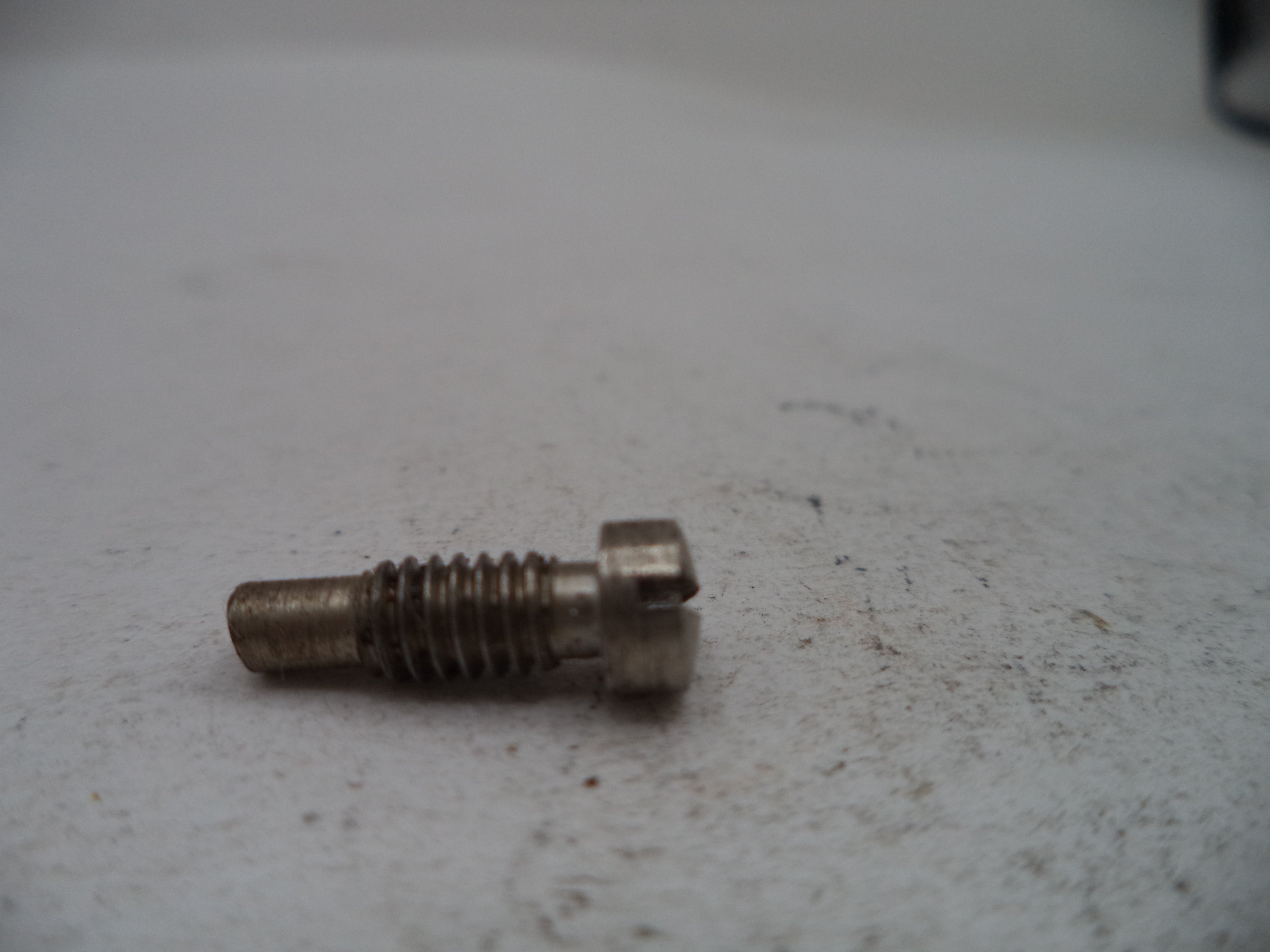 A small metal threaded screw or bolt with a slotted head, resting on a light gray textured surface.