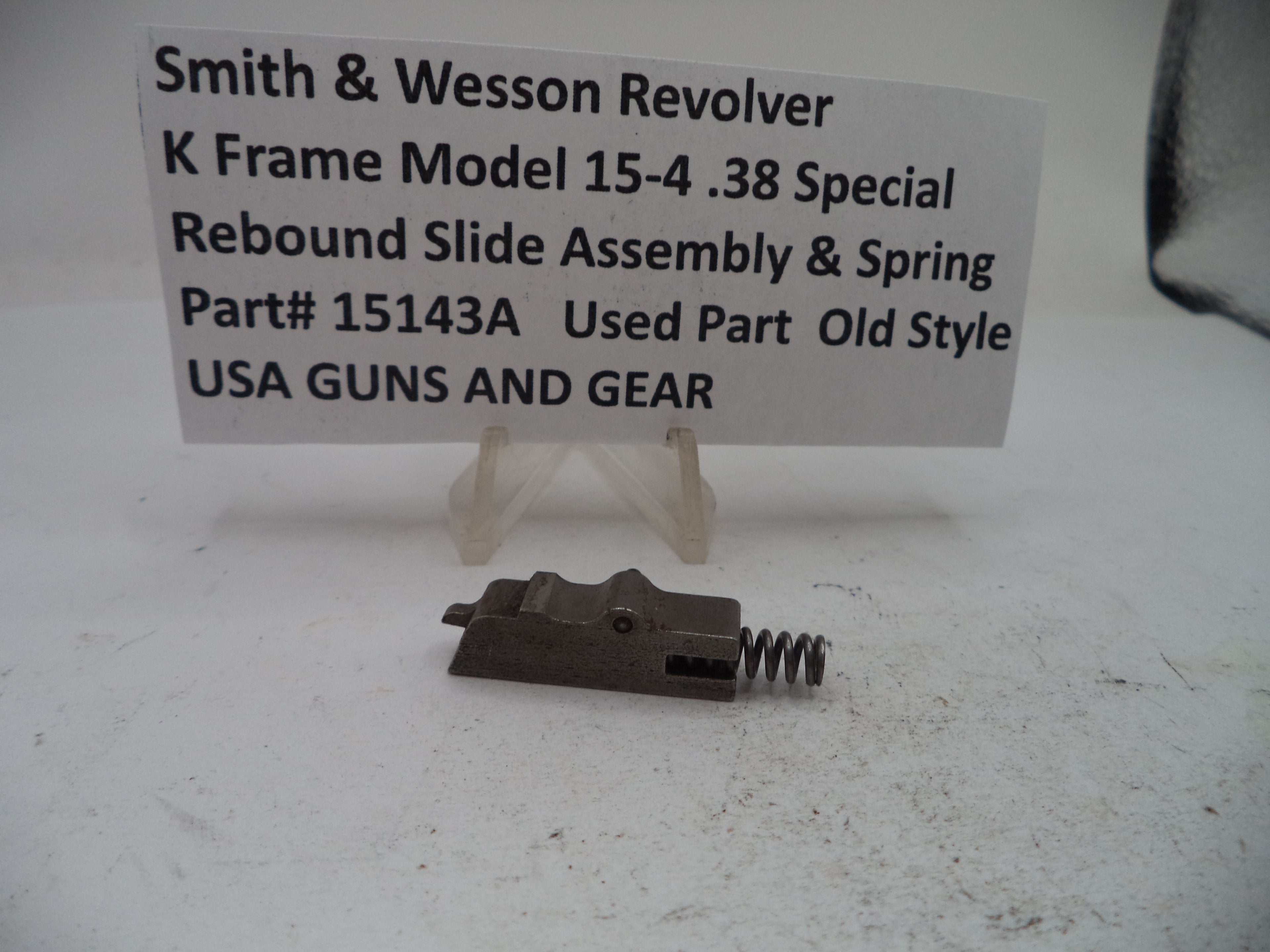 A metal revolver rebound slide assembly with a coil spring, displayed on a small clear stand in front of a white sign that reads 'Smith & Wesson Revolver K Frame Model 15-4 .38 Special Rebound Slide Assembly & Spring Part# 15143A Used Part Old Style USA GUNS AND GEAR'.