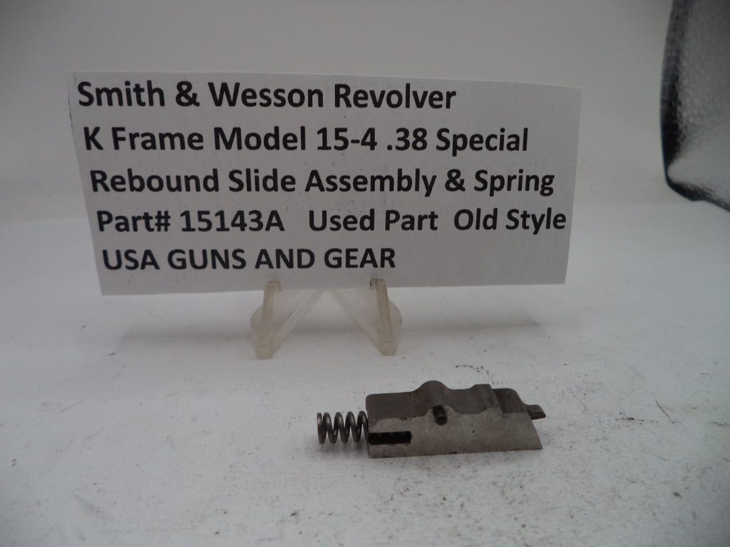 A used metal revolver slide assembly with a coil spring, displayed on a white surface with a white sign reading 'Smith & Wesson Revolver K Frame Model 15-4 .38 Special Rebound Slide Assembly & Spring Part# 15143A Used Part Old Style USA GUNS AND GEAR'.