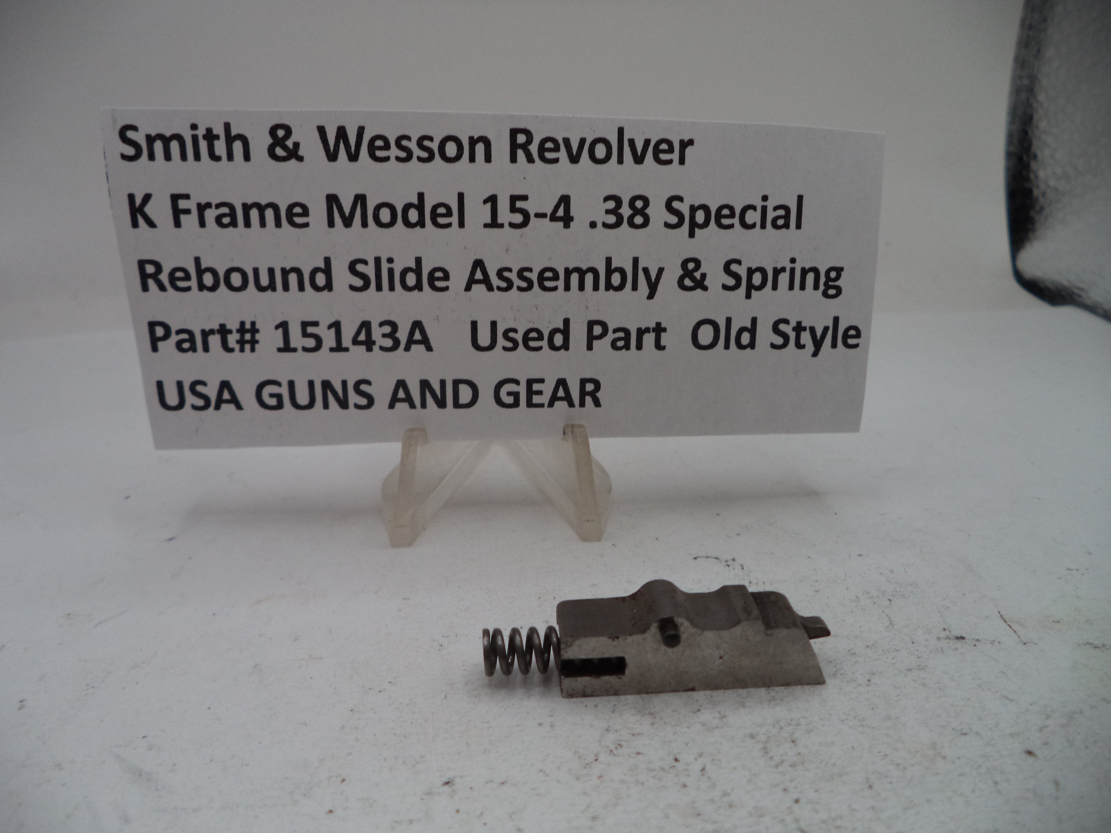 A used metal revolver slide assembly with a coil spring, displayed on a white surface with a white sign reading 'Smith & Wesson Revolver K Frame Model 15-4 .38 Special Rebound Slide Assembly & Spring Part# 15143A Used Part Old Style USA GUNS AND GEAR'.
