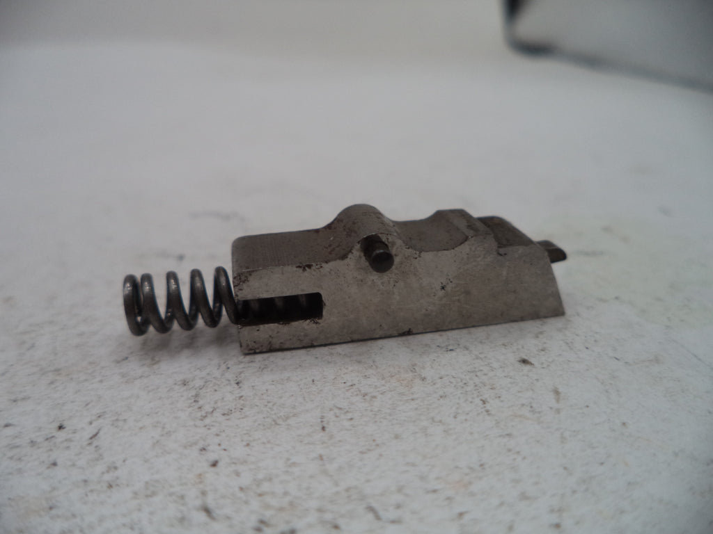 A small metal mechanical component with a coiled spring, photographed on a light textured surface.