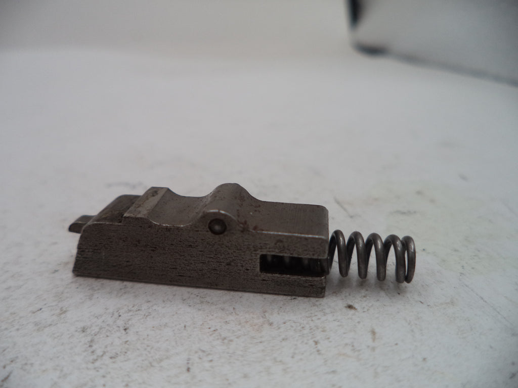 A small metal mechanical component with a dark metallic finish and a coiled spring, photographed on a light off-white surface.