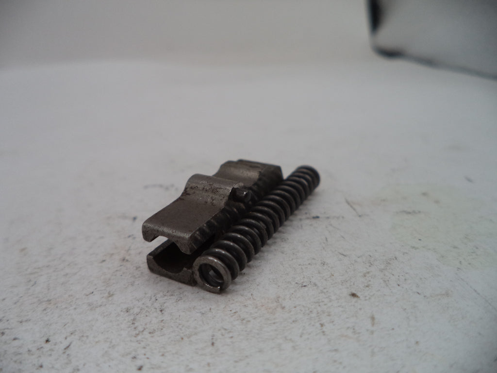 A small dark metal mechanical component with a coiled spring, photographed on a light off-white surface.