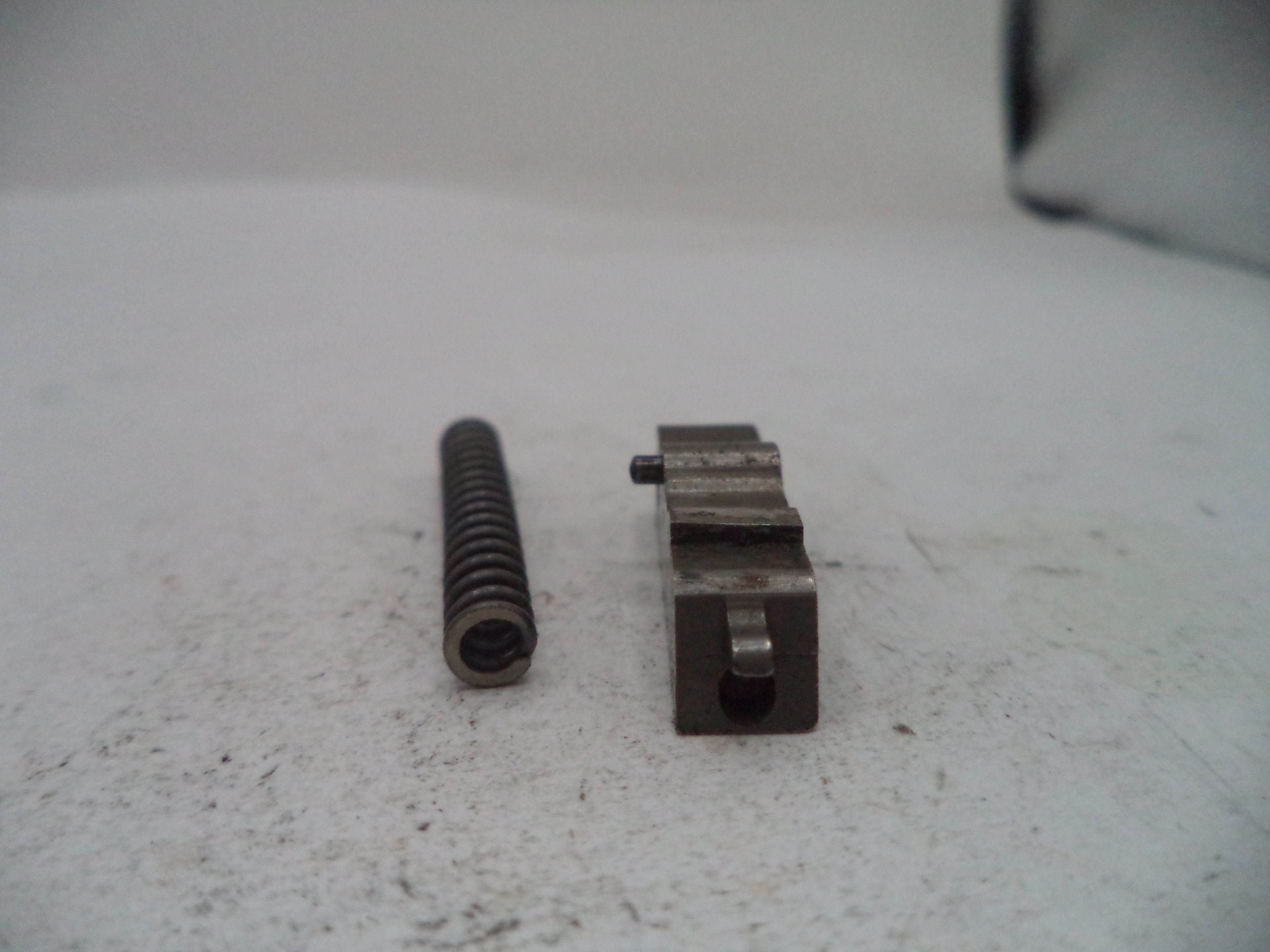 A small metal mechanical component and a dark metal spring placed on a light gray textured surface.