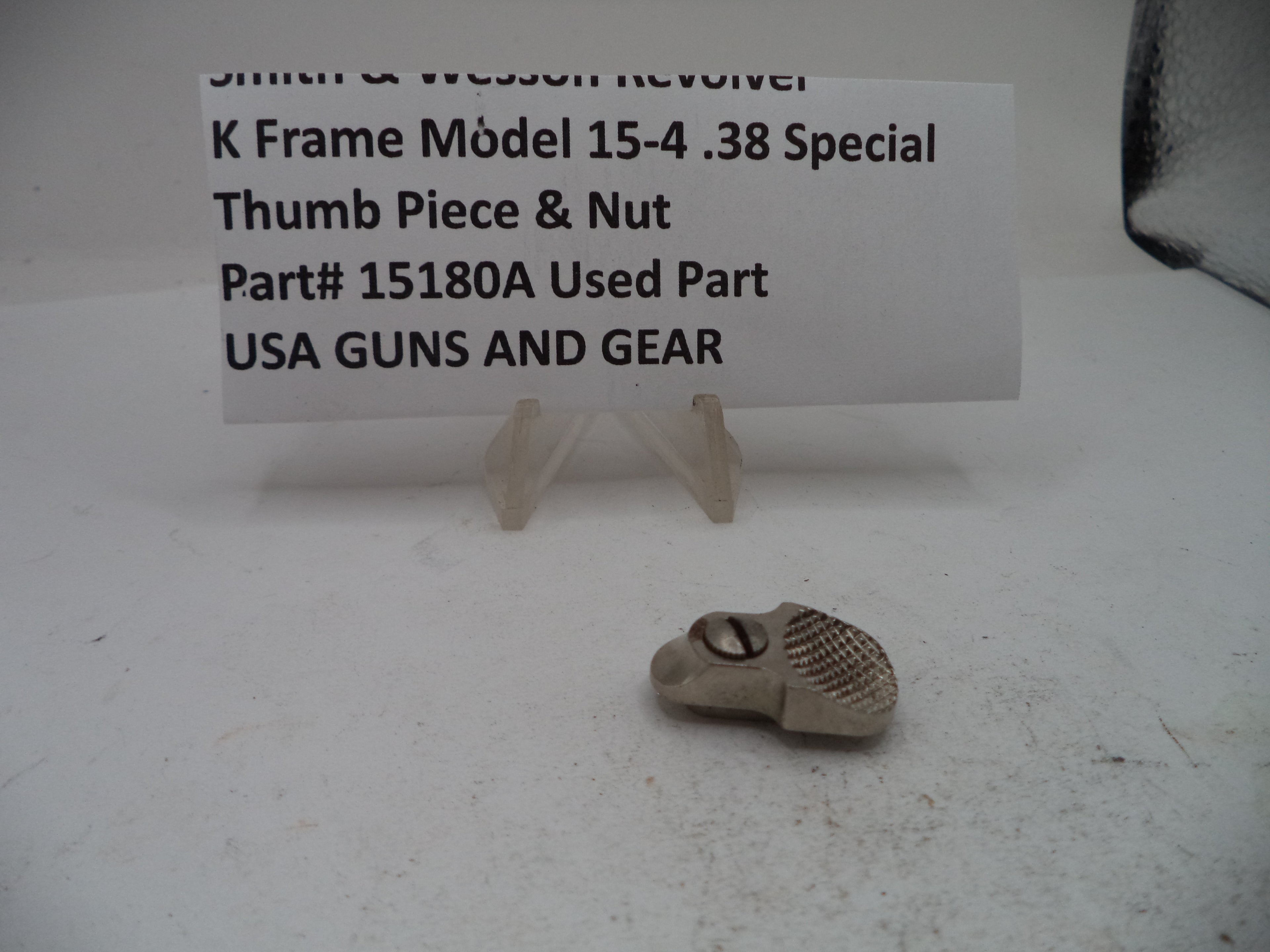 A small metal firearm thumb piece and nut replacement part, part number 15180A, displayed on a white surface in front of a white sign with black text identifying the item.