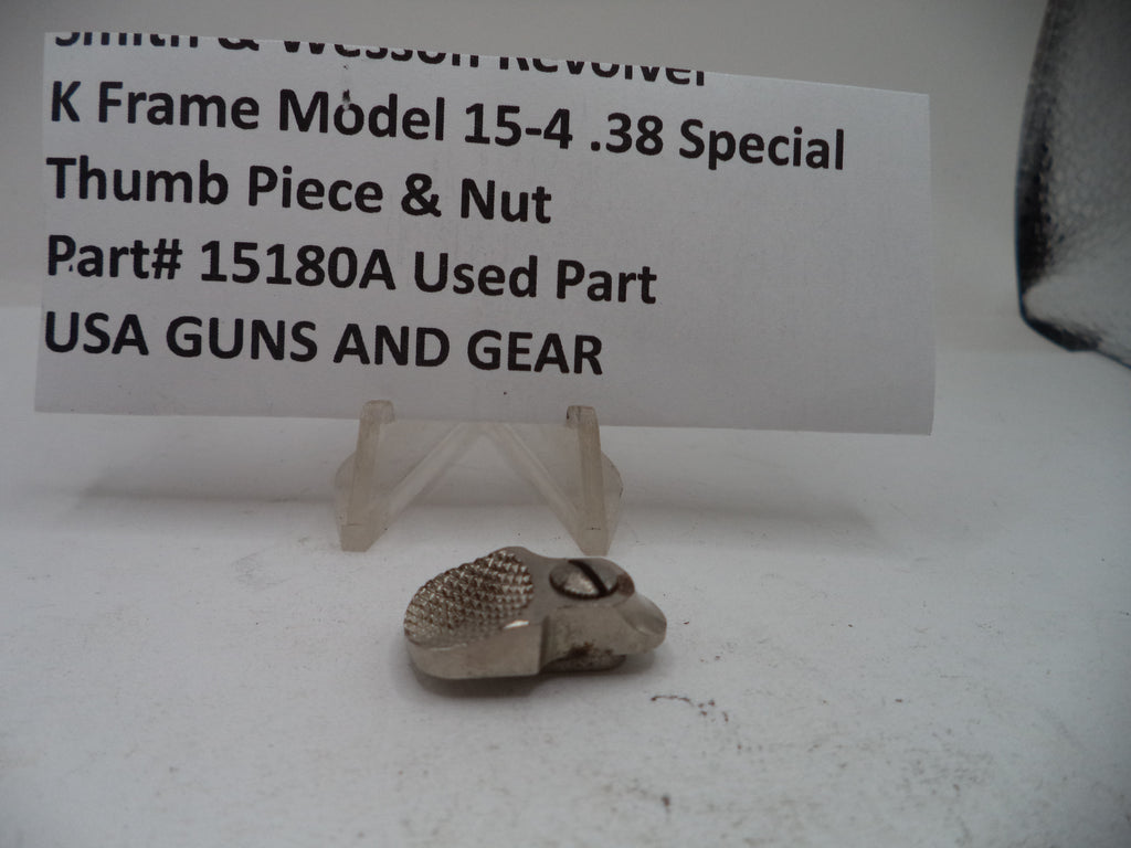 A small used metal thumb piece and nut part for a Smith & Wesson K-frame revolver, displayed on a clear plastic stand in front of a white paper label card with black text.