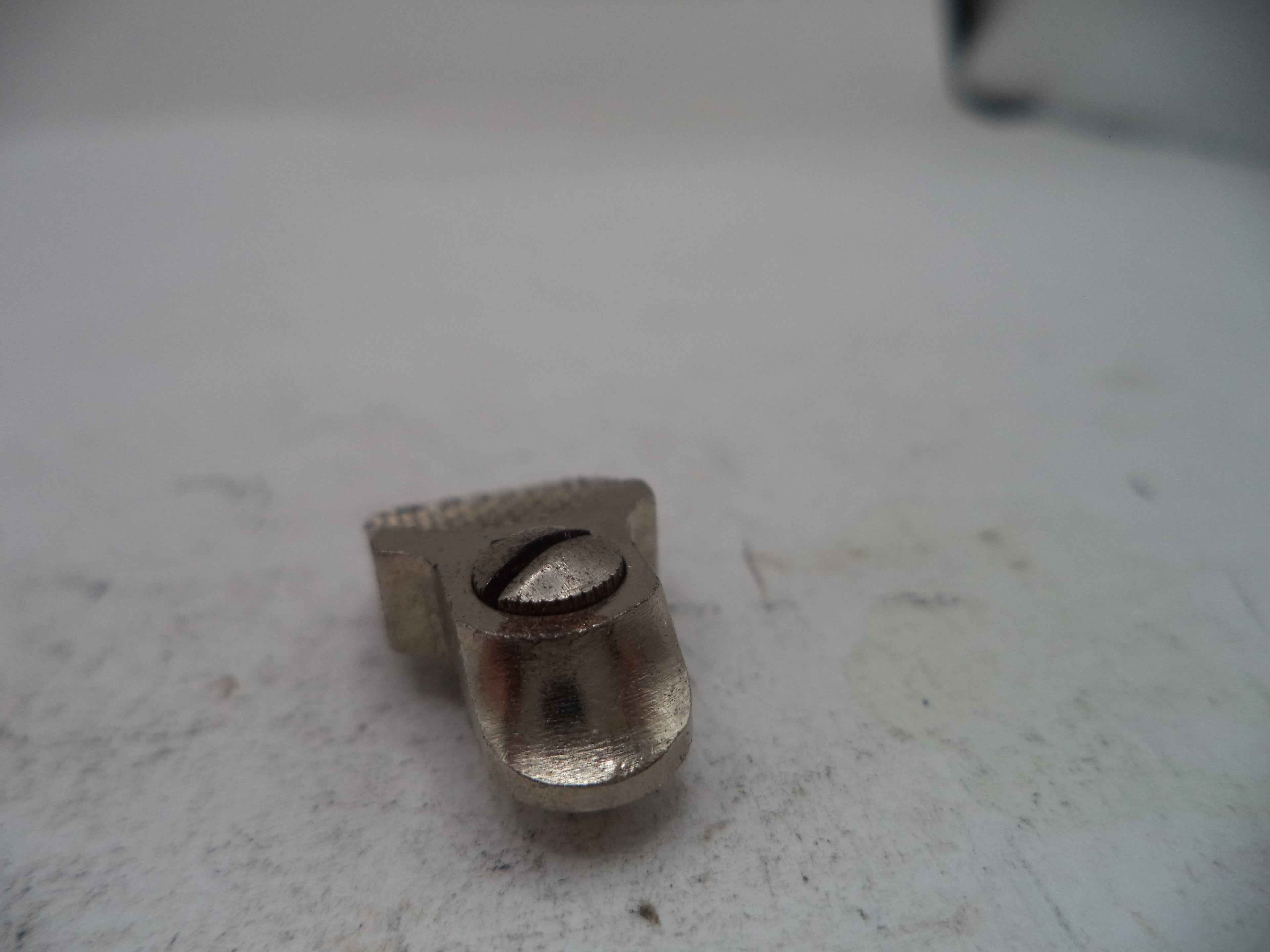 A small metallic silver hardware component, possibly a bracket or mounting piece, photographed on a worn off-white surface.