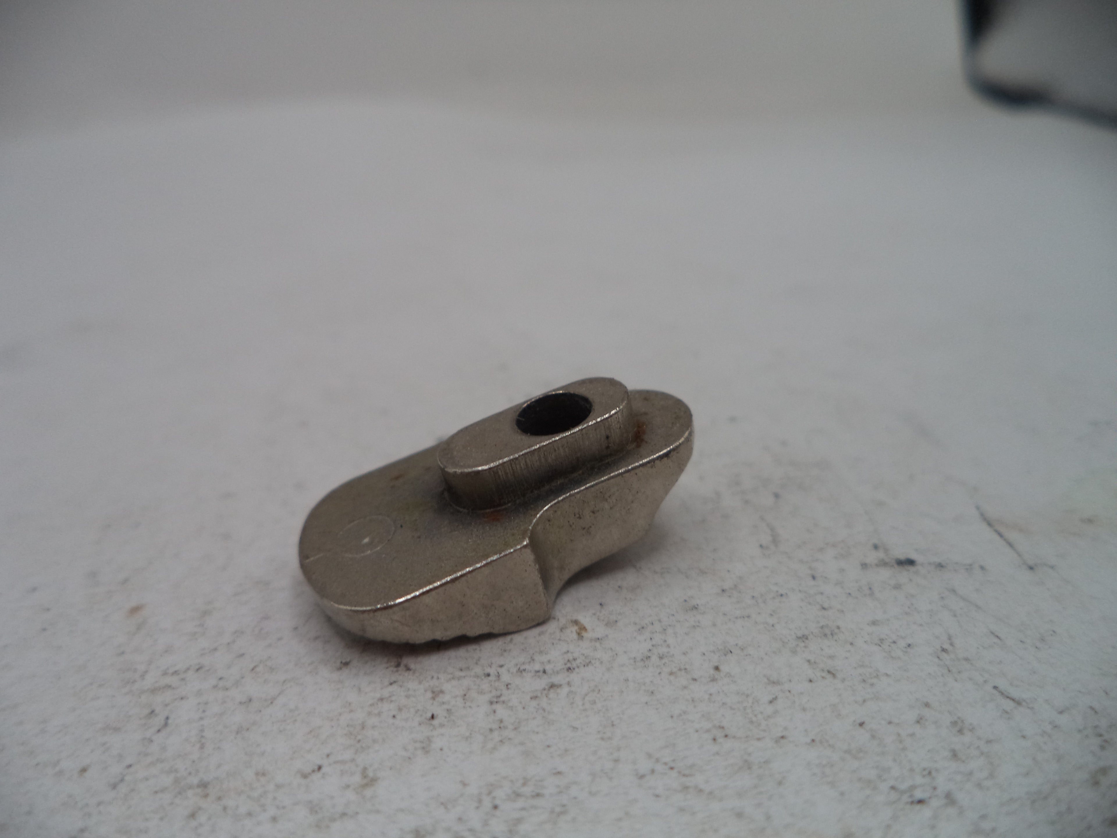 A small metallic silver hardware component or tool piece with a central hole, resting on a textured light gray surface.