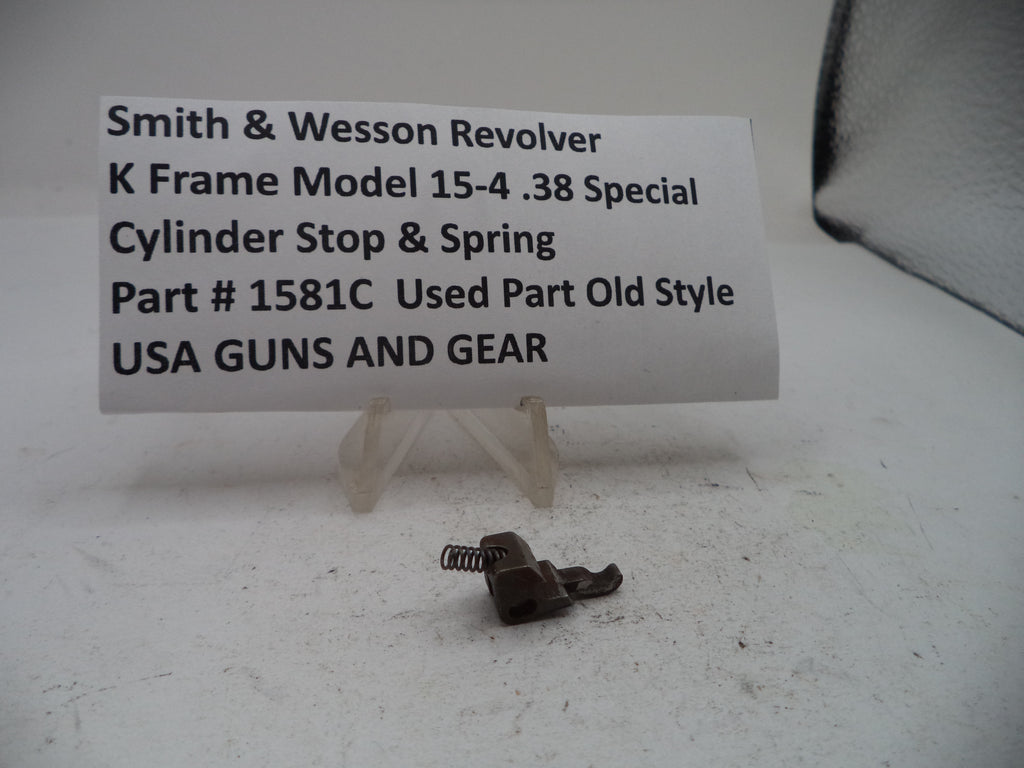 A small metal cylinder stop and spring part displayed on a white surface in front of a white sign that reads 'Smith & Wesson Revolver K Frame Model 15-4 .38 Special Cylinder Stop & Spring Part # 1581C Used Part Old Style USA GUNS AND GEAR'.