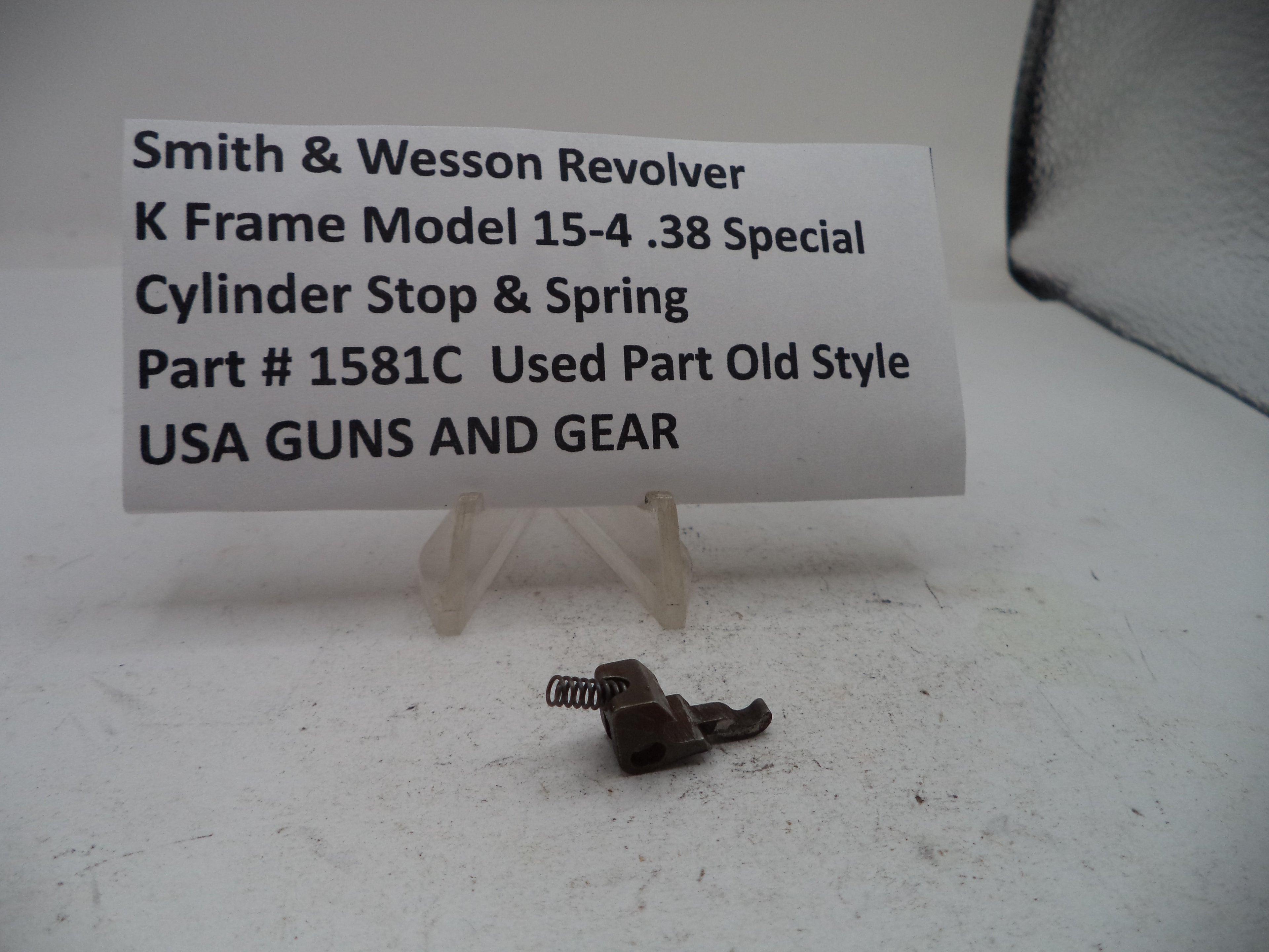 A small metal cylinder stop and spring part displayed on a white surface in front of a white sign that reads 'Smith & Wesson Revolver K Frame Model 15-4 .38 Special Cylinder Stop & Spring Part # 1581C Used Part Old Style USA GUNS AND GEAR'.
