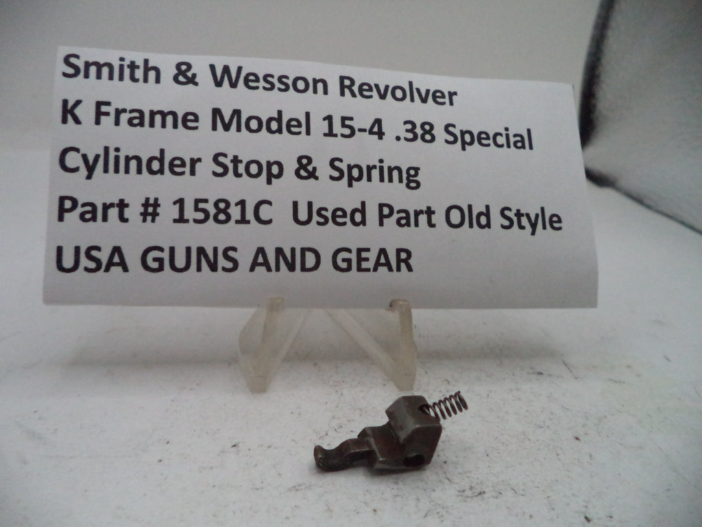 A small metal cylinder stop and spring part displayed on a clear stand in front of a white label card that reads 'Smith & Wesson Revolver K Frame Model 15-4 .38 Special Cylinder Stop & Spring Part # 1581C Used Part Old Style USA GUNS AND GEAR'.
