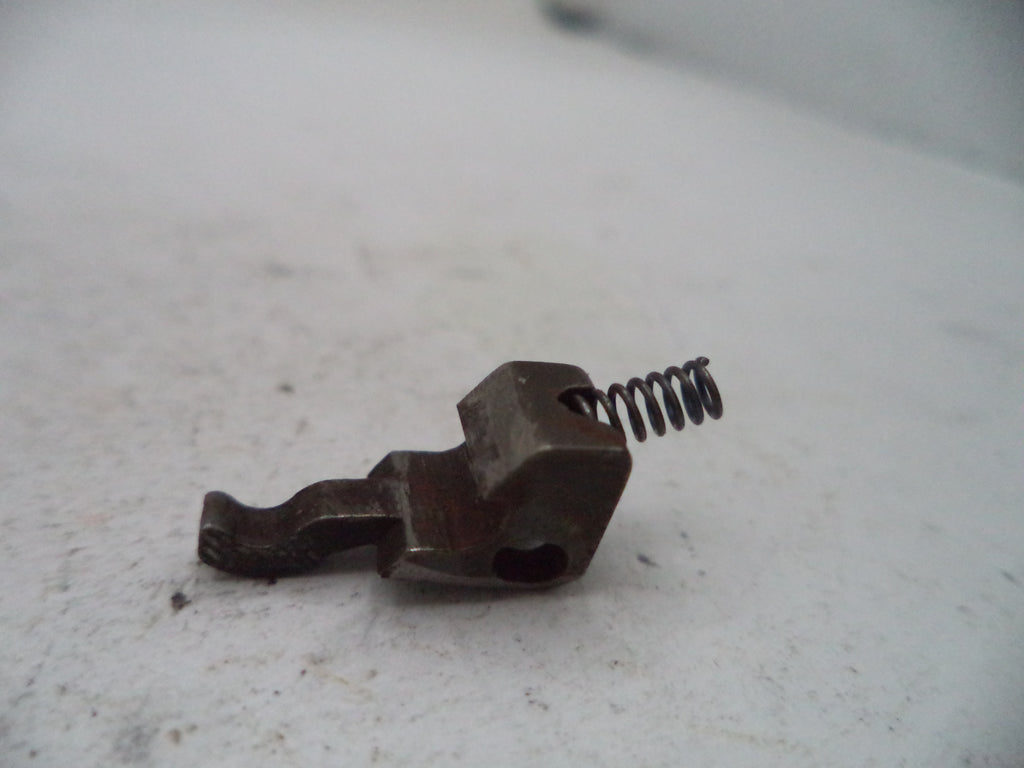 A small dark metal mechanical component with a coiled spring, photographed on a light off-white surface.
