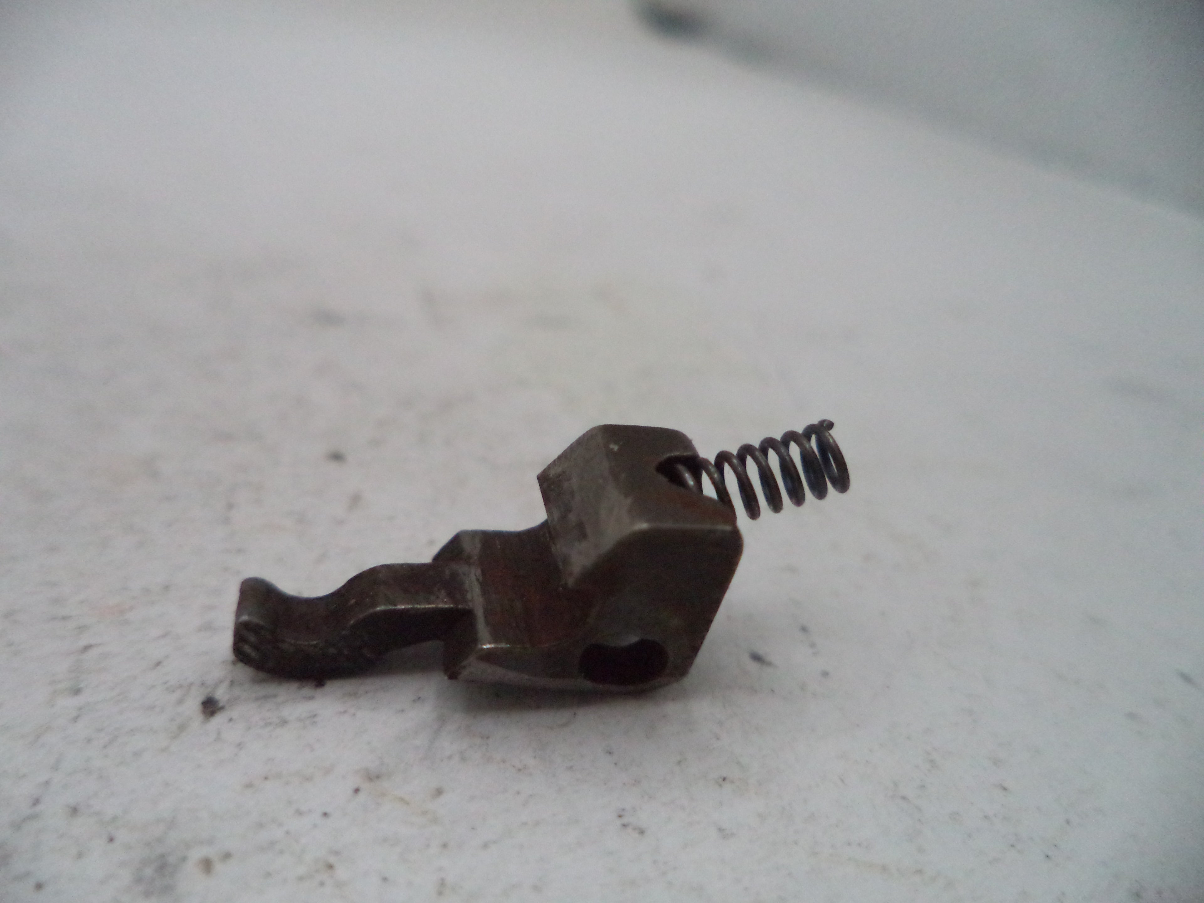 A small dark metal mechanical component with a coiled spring, photographed on a light off-white surface.
