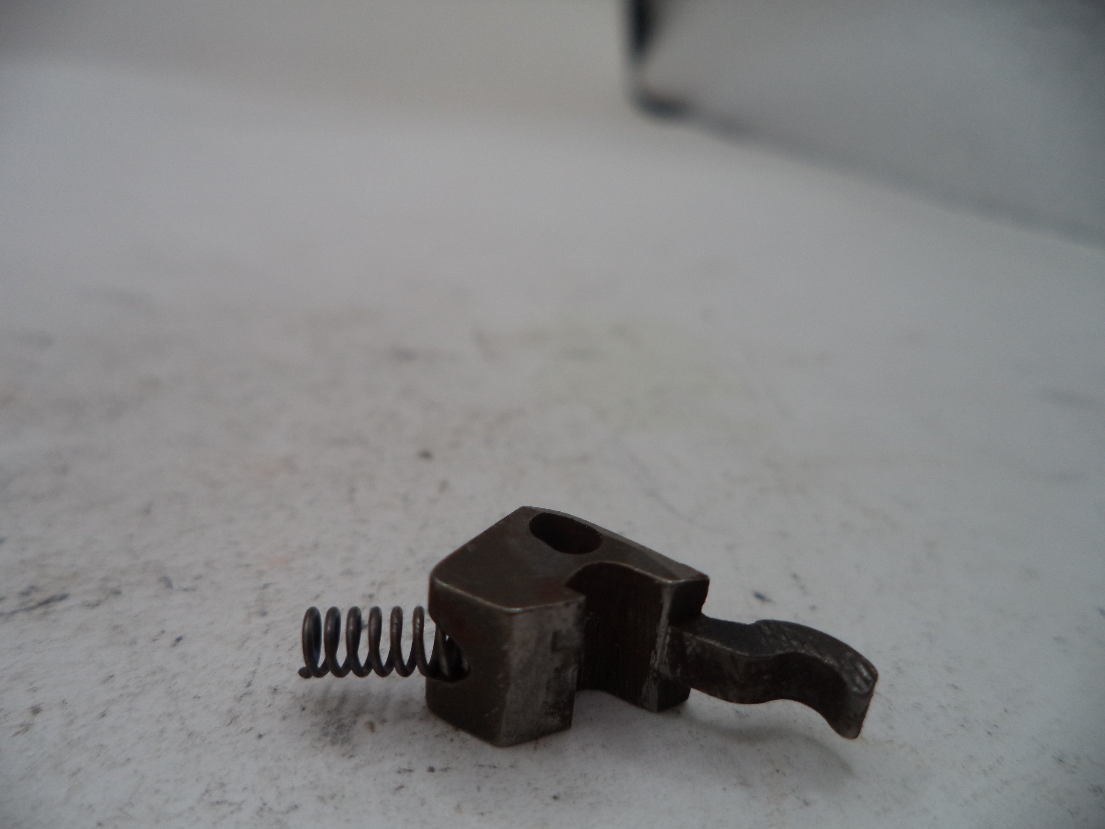 A small dark metal mechanical component with a coiled spring, photographed on a light gray surface.