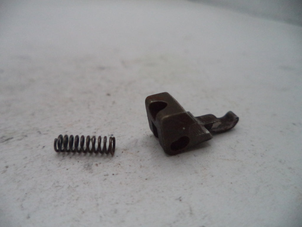 A small metal mechanical component with a dark brown oxidized finish and a separate coiled spring, photographed on a light gray textured surface.