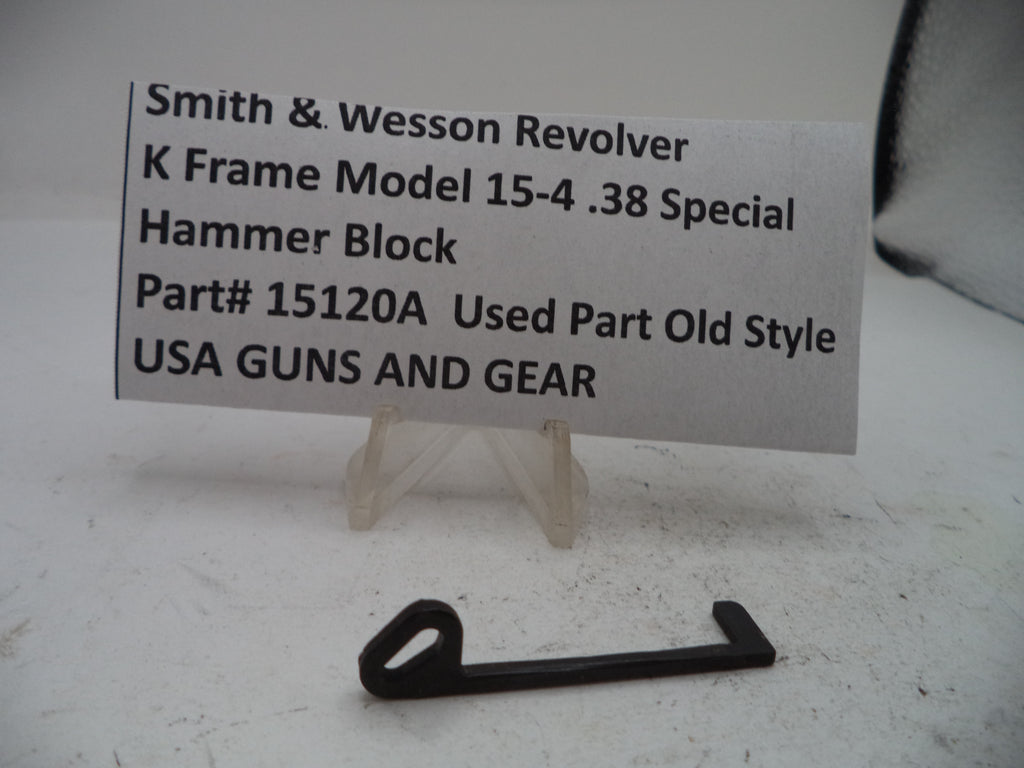 A small black metal hammer block part displayed on a clear stand in front of a printed label card that reads 'Smith & Wesson Revolver K Frame Model 15-4 .38 Special Hammer Block Part# 15120A Used Part Old Style USA GUNS AND GEAR.