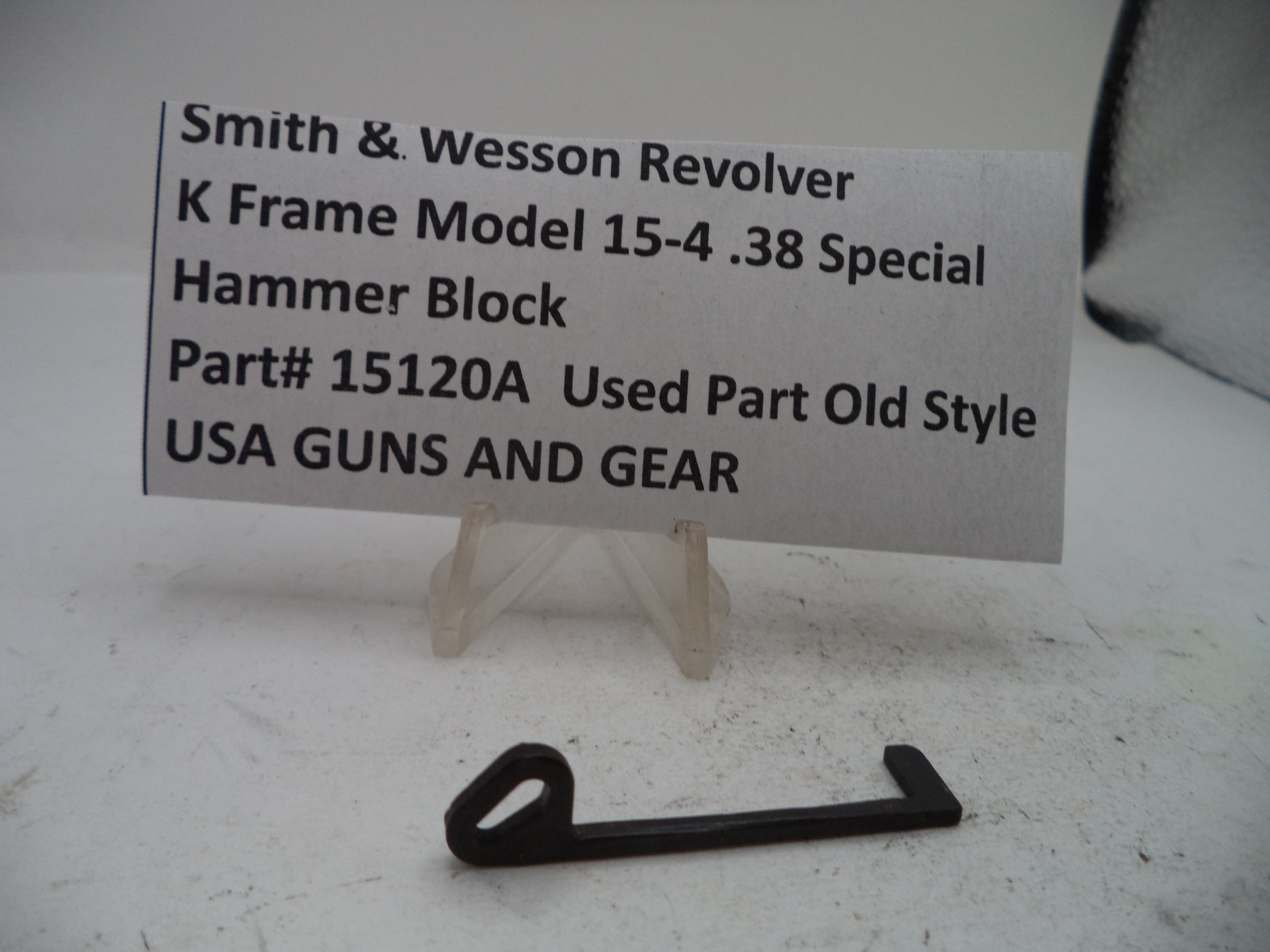 A small black metal hammer block part displayed on a clear stand in front of a printed label card that reads 'Smith & Wesson Revolver K Frame Model 15-4 .38 Special Hammer Block Part# 15120A Used Part Old Style USA GUNS AND GEAR.