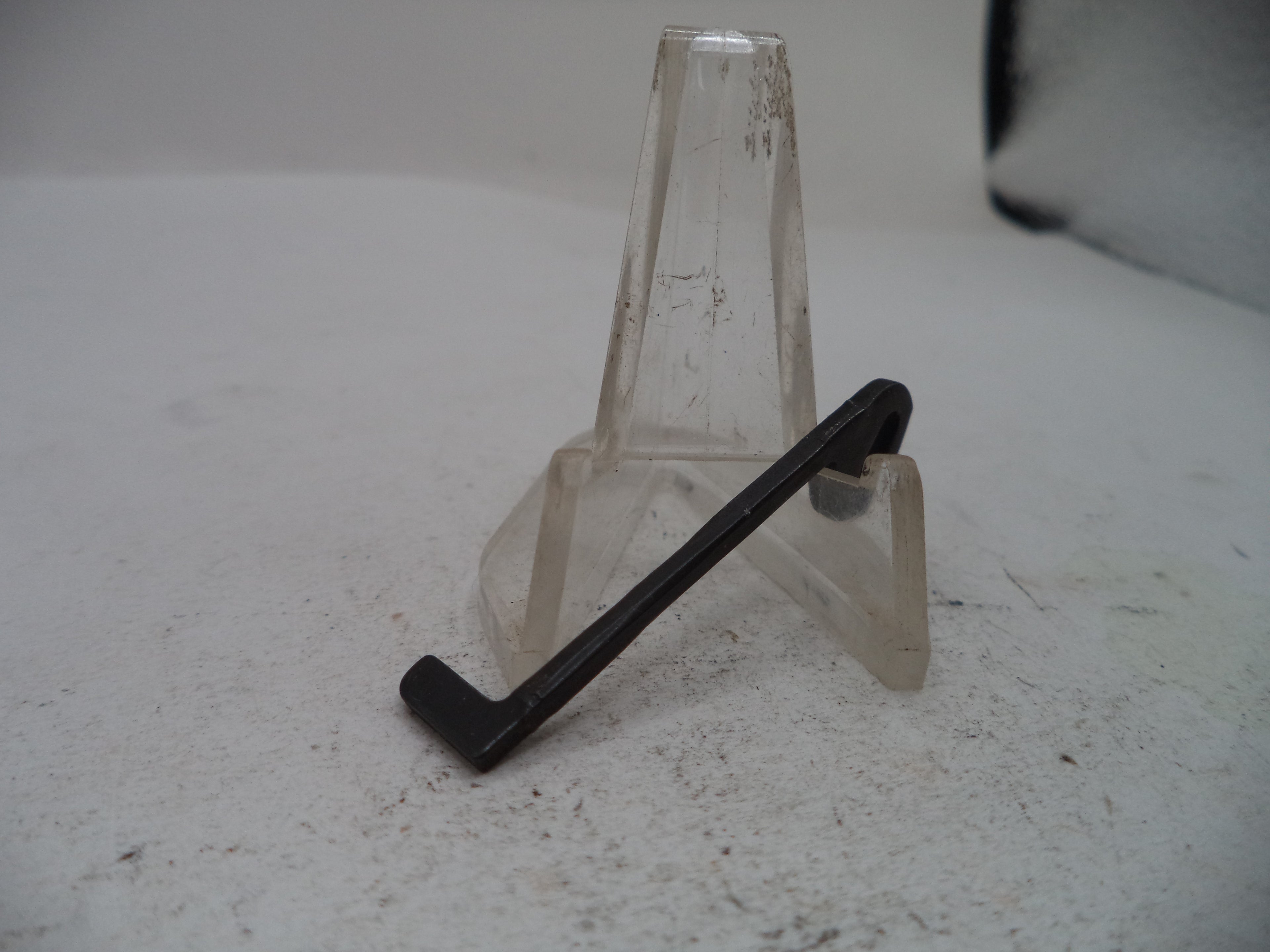 A black metal L-shaped bracket or clip displayed on a clear plastic stand against a light gray background.