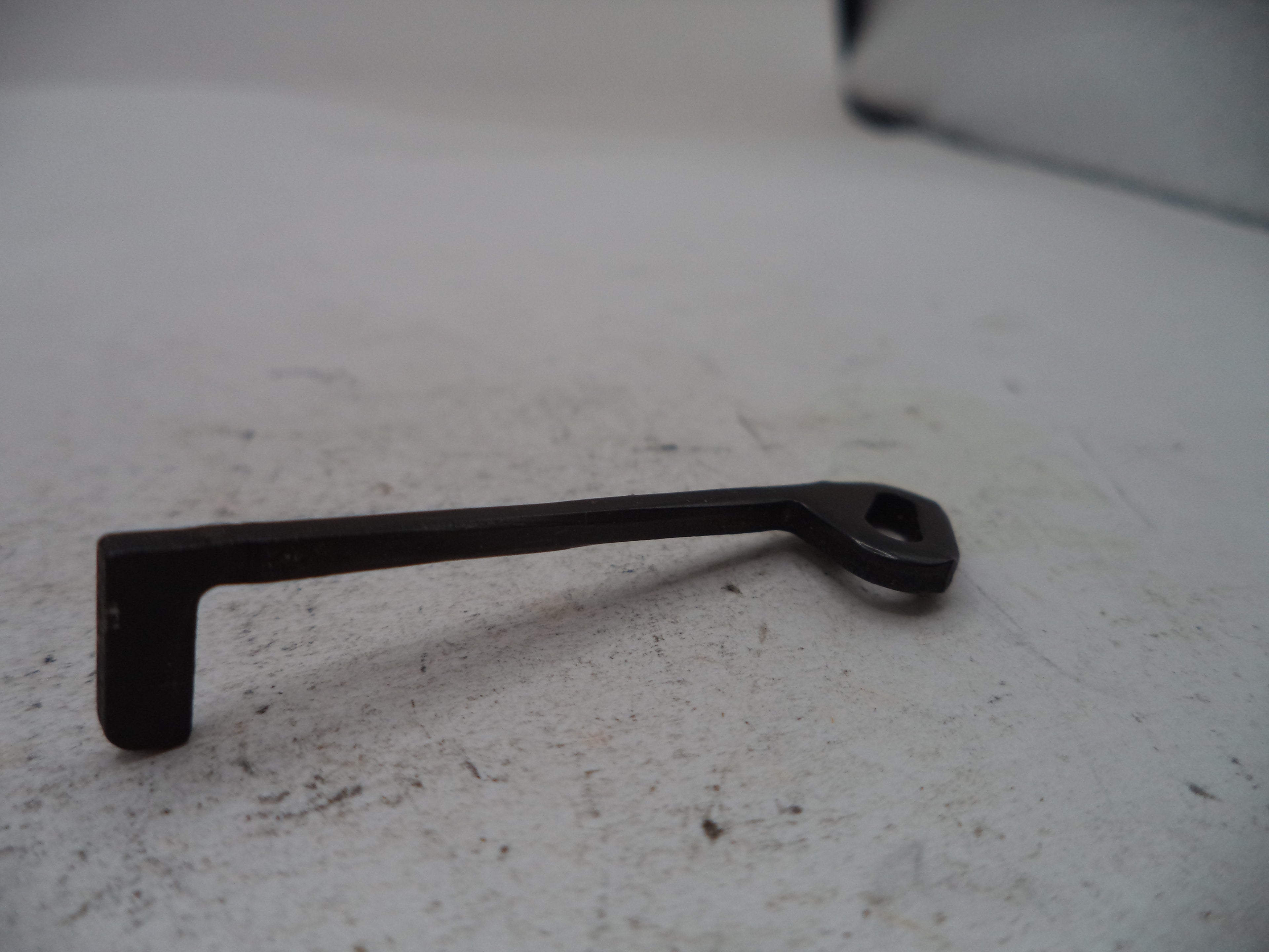 A small black metal hardware piece with a bent L-shaped end and a looped handle, photographed on a light gray surface.