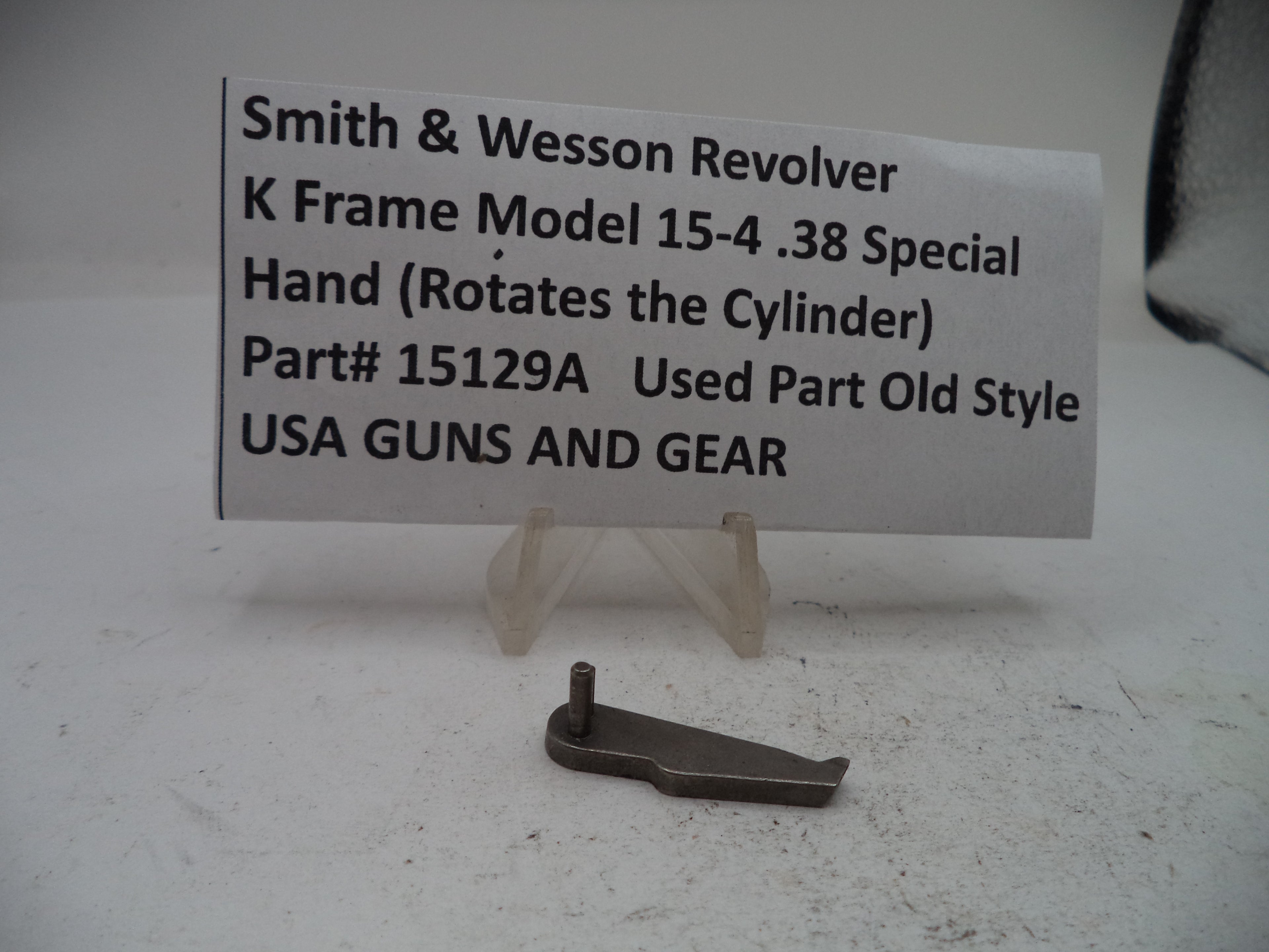A small metal revolver part labeled 'Smith & Wesson Revolver K Frame Model 15-4 .38 Special Hand (Rotates the Cylinder)' on a white display stand in front of a paper sign.