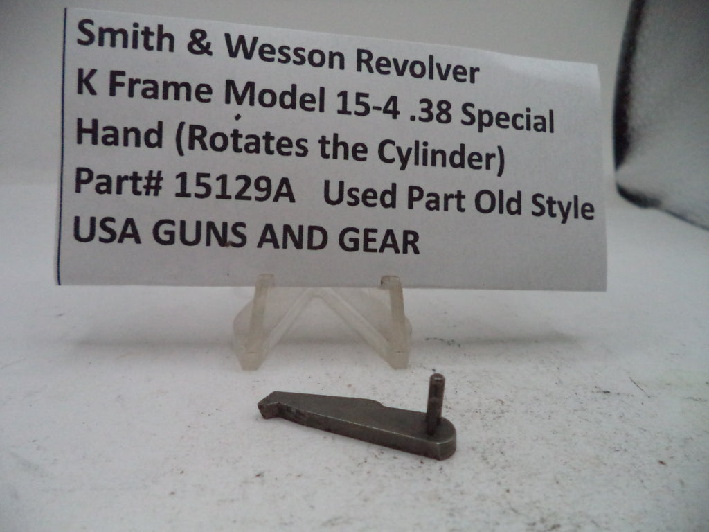 A small metal revolver part displayed on a clear plastic stand in front of a white sign that reads 'Smith & Wesson Revolver K Frame Model 15-4 .38 Special Hand (Rotates the Cylinder) Part# 15129A Used Part Old Style USA GUNS AND GEAR'.