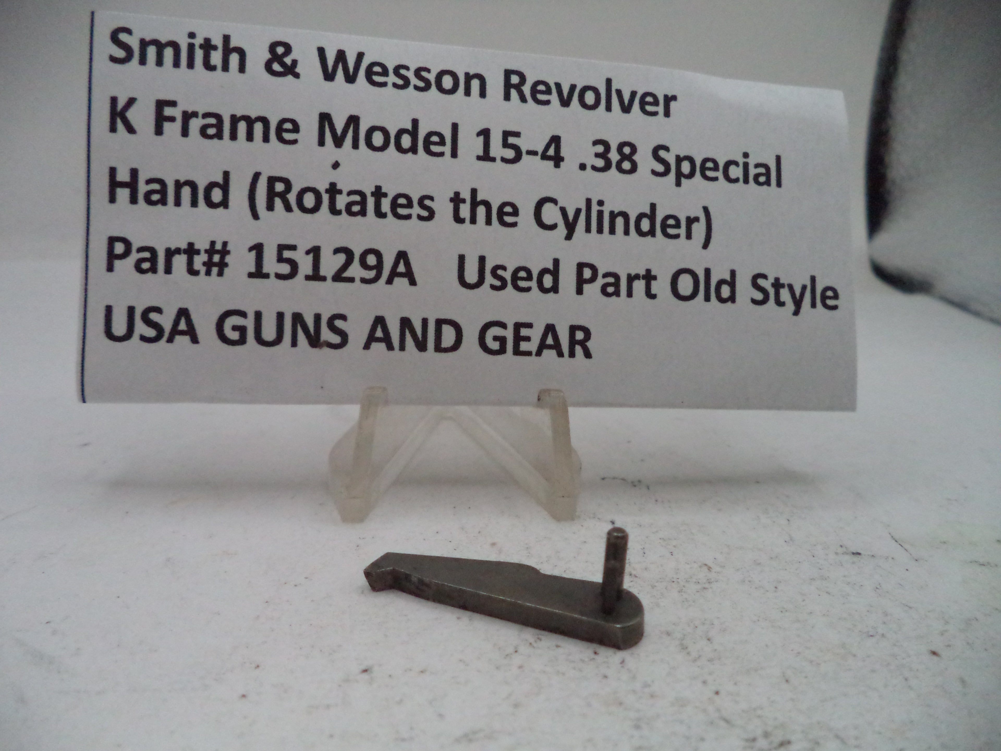 A small metal revolver part displayed on a clear plastic stand in front of a white sign that reads 'Smith & Wesson Revolver K Frame Model 15-4 .38 Special Hand (Rotates the Cylinder) Part# 15129A Used Part Old Style USA GUNS AND GEAR'.