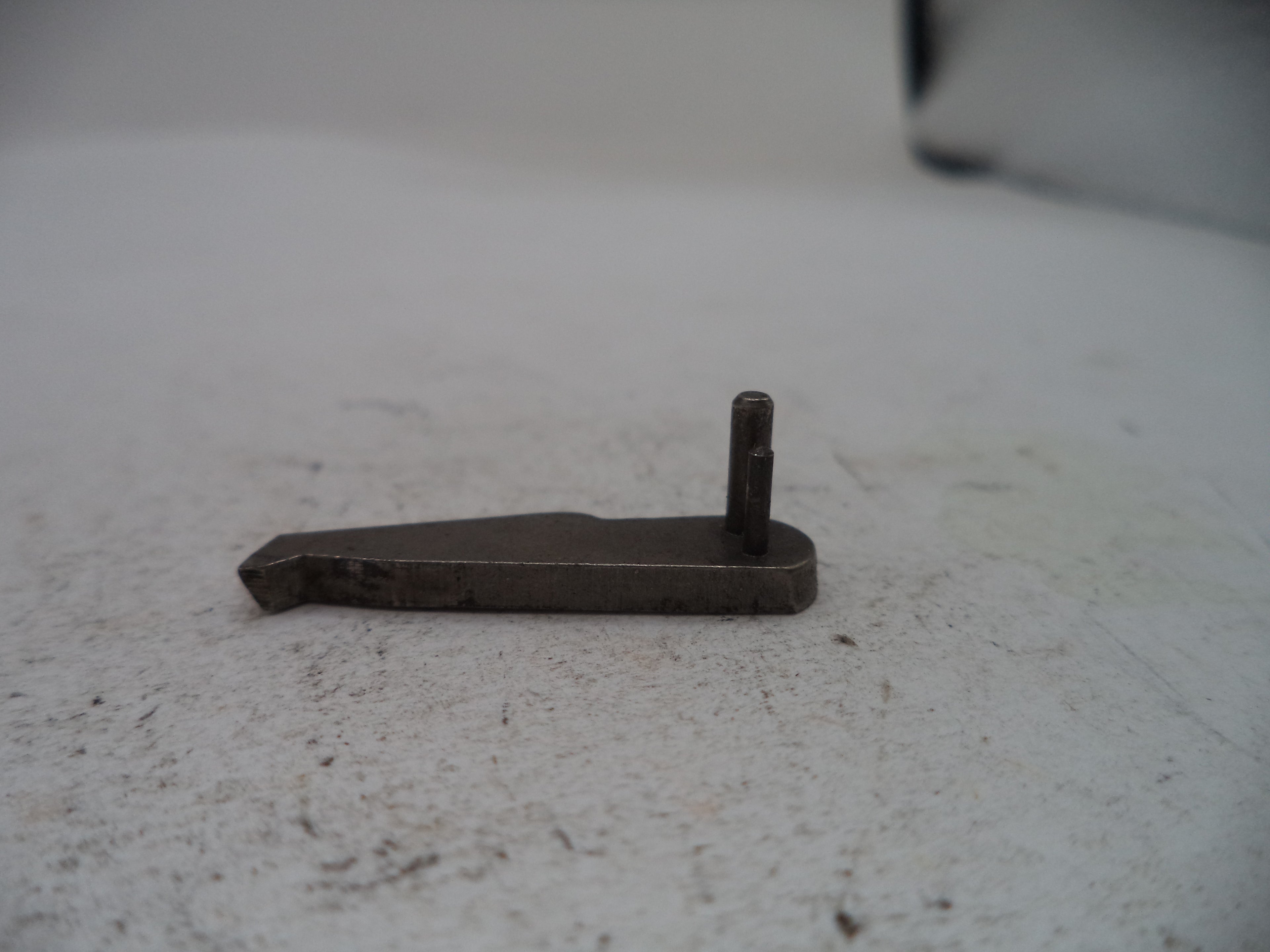 A small dark metal hardware component with a flat rectangular plate and a short vertical pin, photographed on a light textured surface.