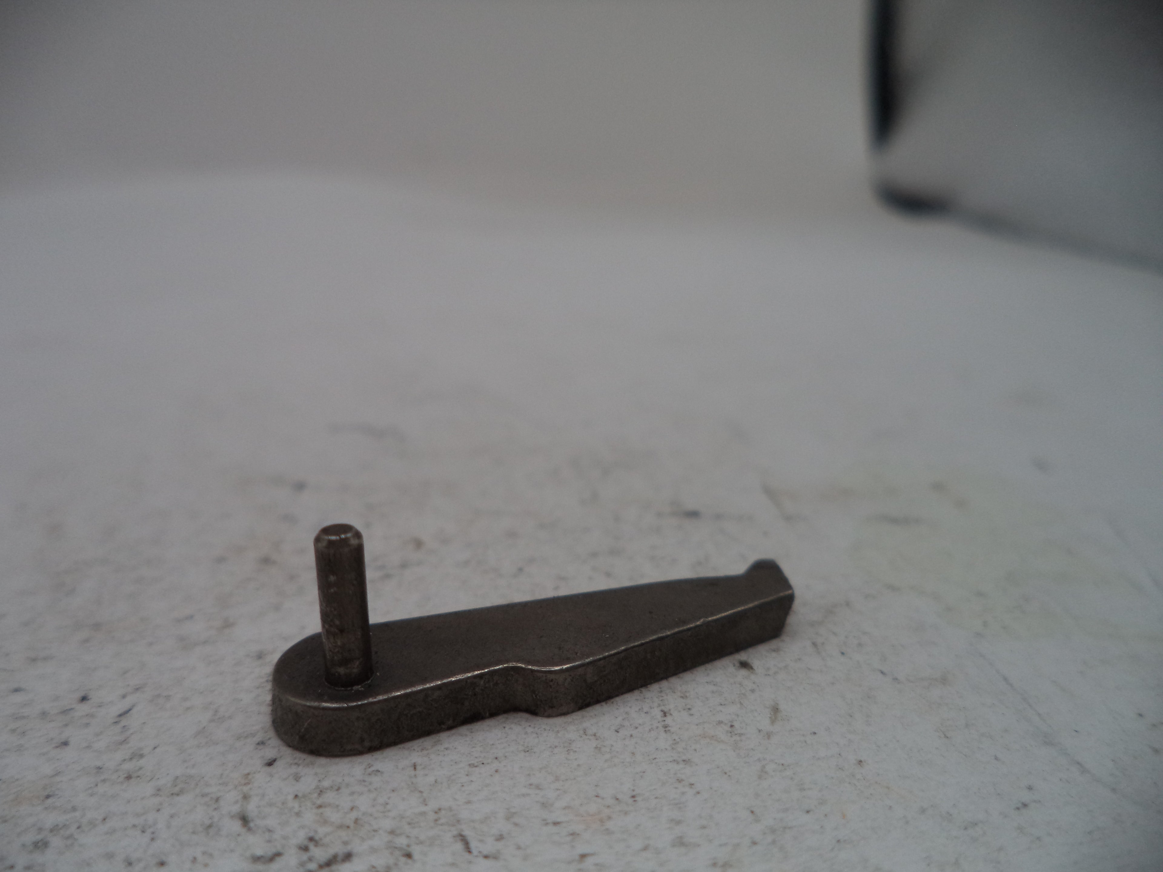 A small dark metal hardware piece with a flat triangular blade and a short vertical pin, photographed on a light gray textured surface.