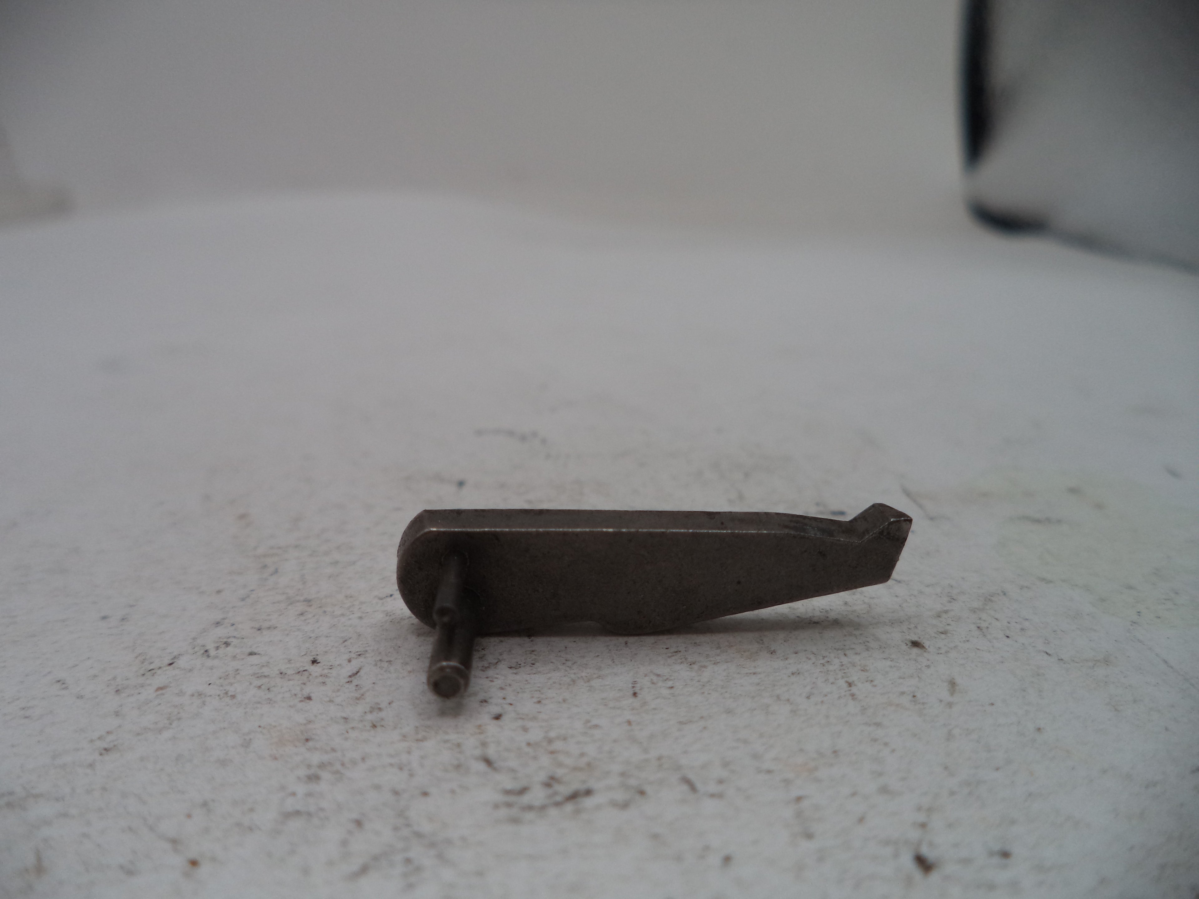 A small dark metal hardware piece with a flat rectangular body and a short pin extending downward, photographed on a light off-white surface.