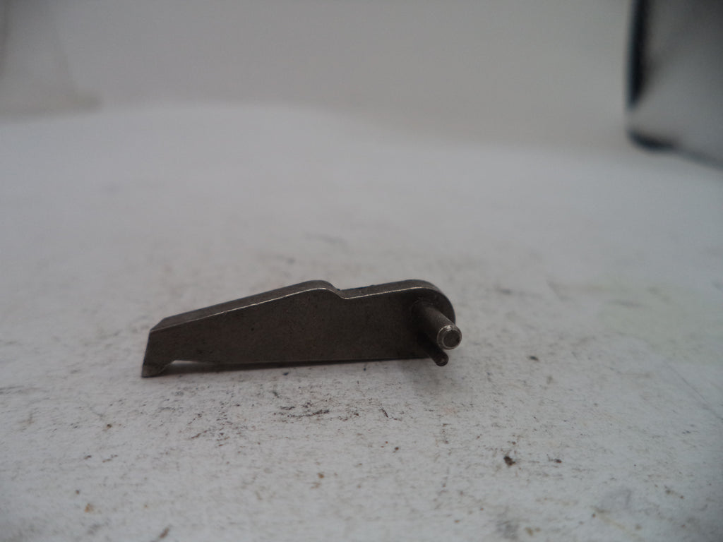 A small dark metal mechanical component with a flat angled body and a protruding pin, photographed on a light gray textured surface.