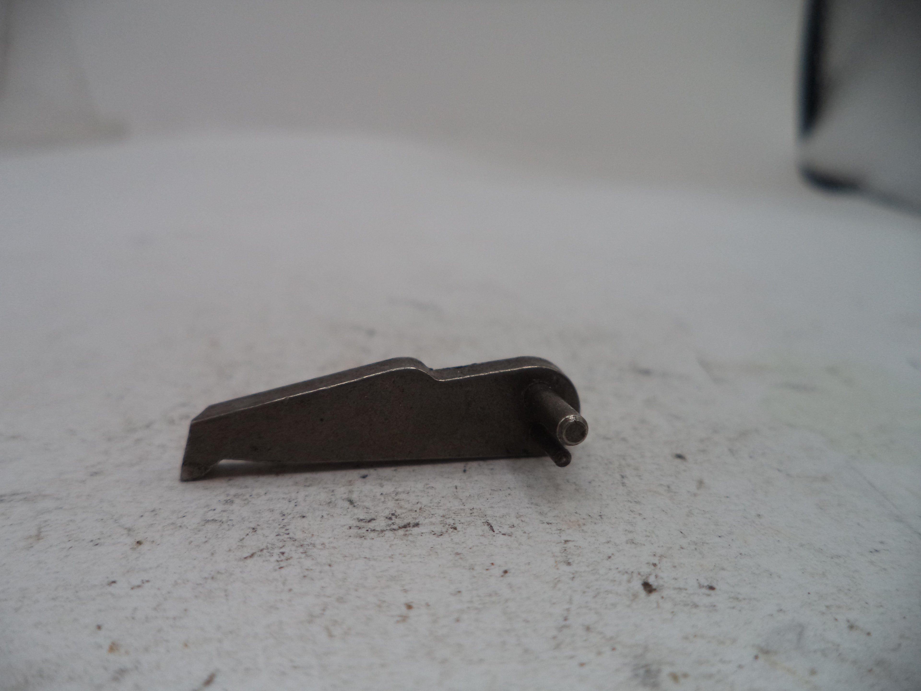 A small dark metal mechanical component with a flat angled body and a protruding pin, photographed on a light gray textured surface.