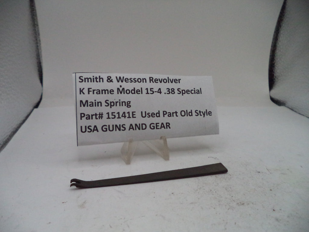 A small metal part labeled 'Smith & Wesson Revolver K Frame Model 15-4 .38 Special Main Spring Part# 15141E Used Part Old Style USA GUNS AND GEAR' displayed on a small stand against a plain light background.