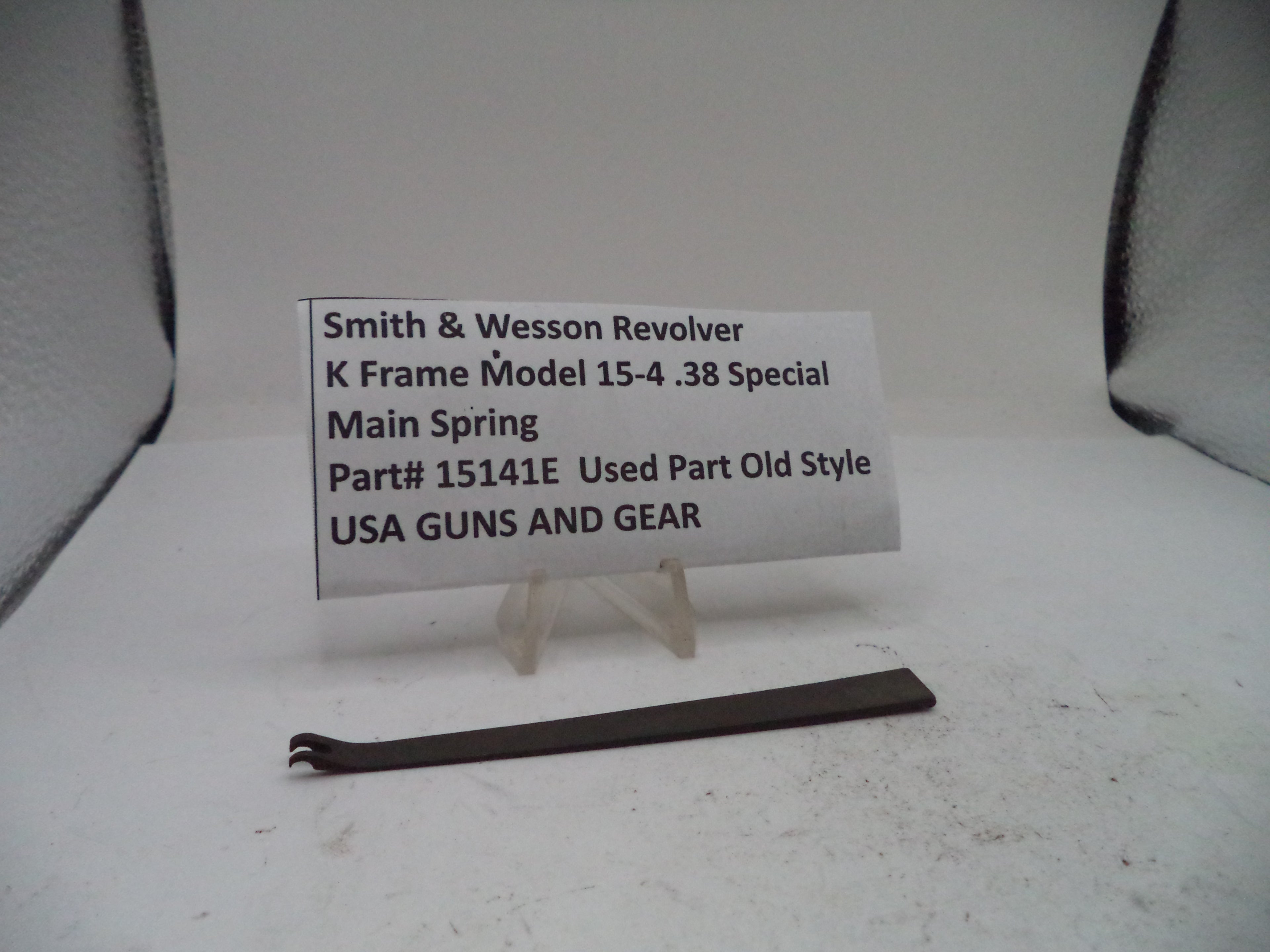 A small metal part labeled 'Smith & Wesson Revolver K Frame Model 15-4 .38 Special Main Spring Part# 15141E Used Part Old Style USA GUNS AND GEAR' displayed on a small stand against a plain light background.